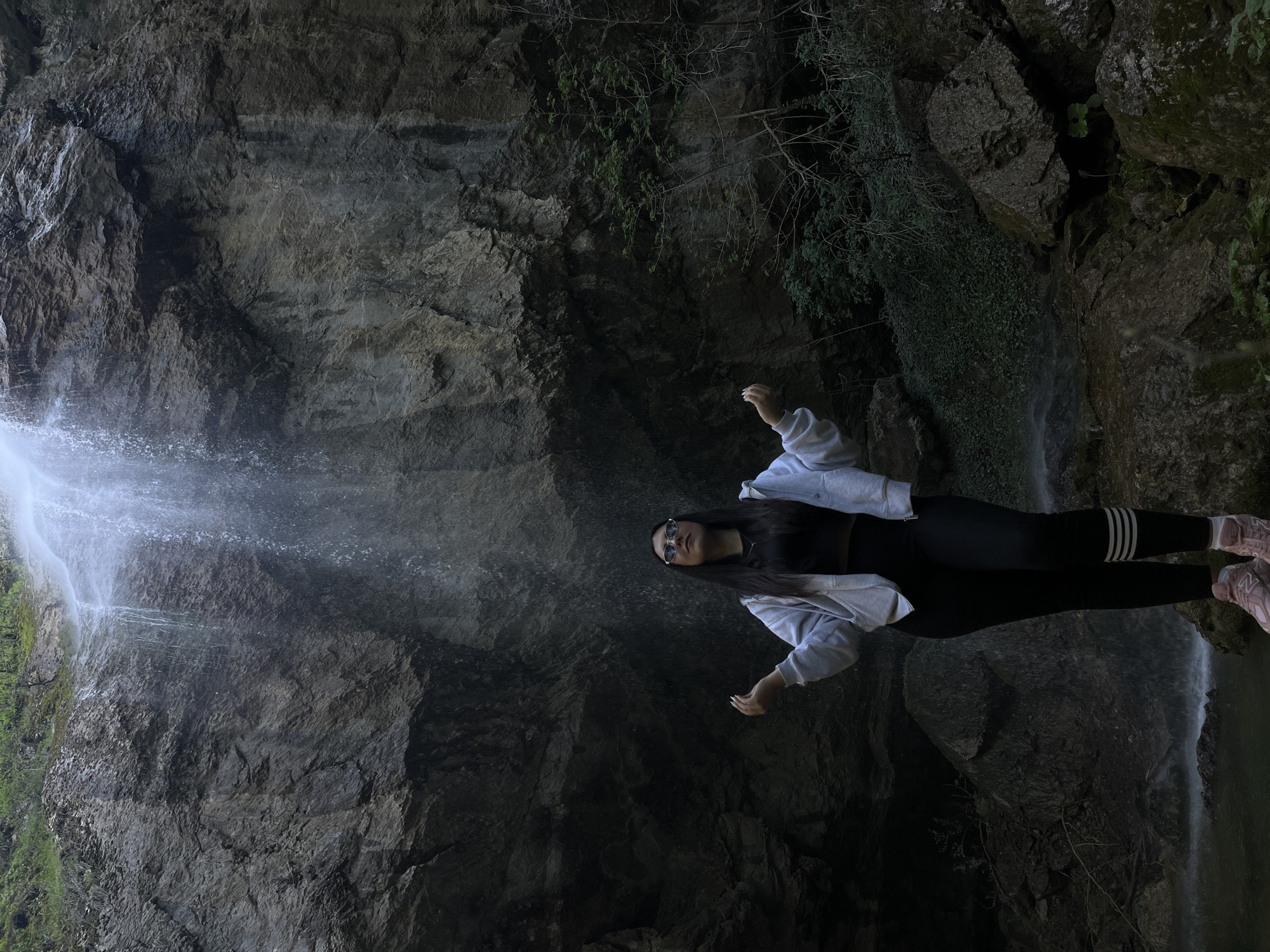 A young woman stands in front of a waterfall in Aleksandrovo, Bulgaria. The woman is wearing black leggings with white stripes down the sides, a black top, a light-colored jacket, and pink sneakers. She has long, dark hair and is wearing sunglasses. Her arms are raised slightly. The waterfall cascades down a rocky cliff face covered with some greenery. The environment is dimly lit, suggesting a cave or grotto-like setting.