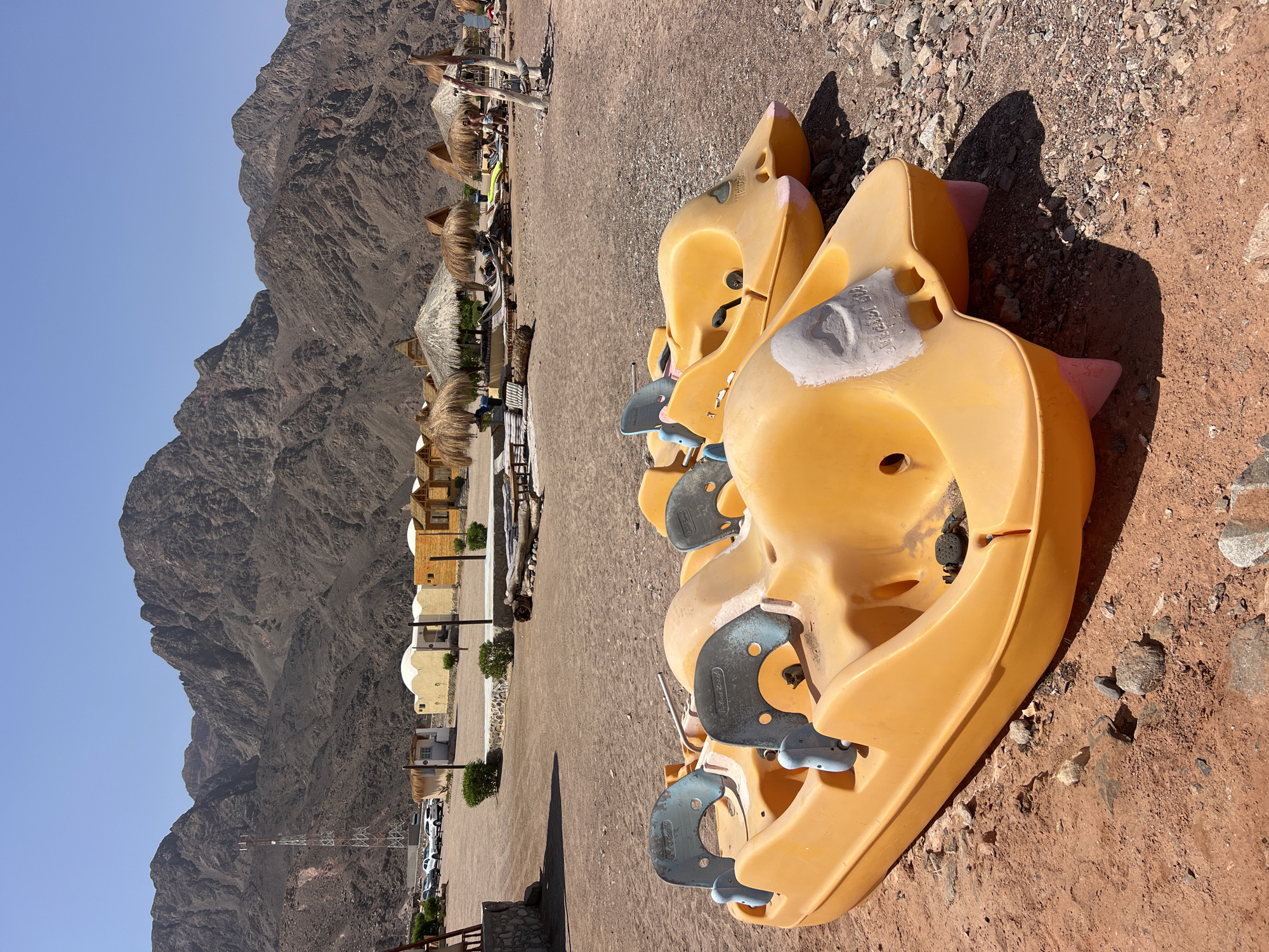 The image shows a beach scene in Nuweiba, Egypt. In the foreground, two bright yellow pedal boats are parked on the sand. The boats are plastic and have blue seats and pedals. Behind the boats, there are several thatched-roof cabanas and some loungers, suggesting a resort or tourist area. In the background, rugged mountains rise against a clear blue sky. The ground is a mix of sand and small rocks. A few people can be seen in the distance, relaxing under the cabanas or walking on the beach. A tall communication tower is visible on the left side of the frame, near some parked cars. The overall impression is a sunny, relaxed atmosphere at a desert beach.