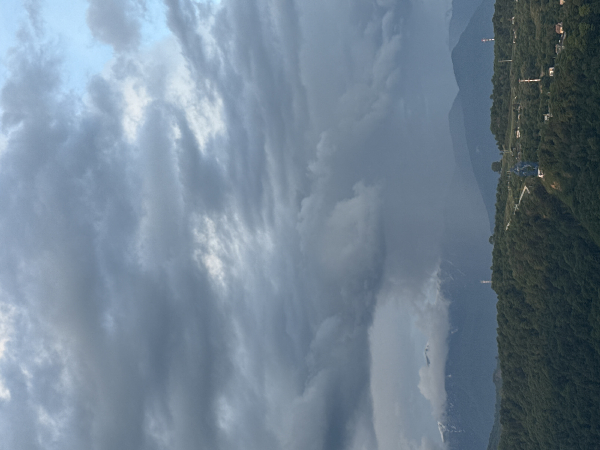 The image shows a mountain landscape under a cloudy sky, likely in Sochi, Russia. The right side of the frame features a densely forested mountain slope with infrastructure elements such as power lines and buildings visible along the ridge. Adjacent to the mountain, a hazy mountain range fades into the background. The sky is overcast with heavy, textured clouds dominating the left side of the image.