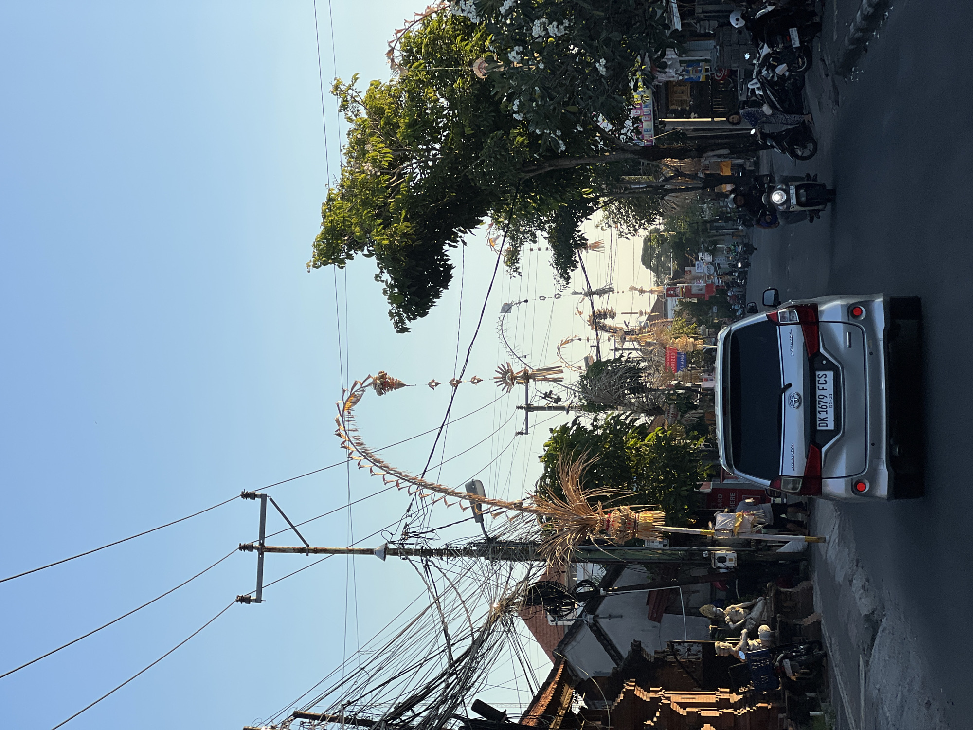 The image depicts a street scene in North Kuta, Indonesia, characterized by its vibrant cultural and urban elements. The photograph is taken from the perspective of a vehicle moving down a paved road, capturing a view of the street as it stretches into the distance. Overhead, a dense network of electrical wires crisscrosses the sky, supported by utility poles, creating a visual tangle that is common in many urban areas.
Lining the sides of the street are buildings that appear to be a mix of shops and residences, with some displaying decorative architectural features and others showing signs of commercial activity. One building on the left features intricate brickwork and appears to house statues displayed outdoors. Along the sidewalk, various items are placed, including motorcycles parked in rows.
A prominent feature of the scene is the presence of traditional Balinese decorations, known as "penjor," which are tall, curved bamboo poles adorned with woven offerings and palm leaves. These are erected along the street, suggesting a cultural or religious event is being celebrated or has recently taken place. The decorations are elaborate and festive, adding a unique cultural dimension to the urban landscape.
The street is active with traffic, including a silver car in the foreground, which is the viewpoint of the photograph, and several motorcycles in motion and parked. People are visible on the sidewalks and near the buildings, engaged in various activities. The overall ambiance is that of a bustling street in a tropical climate, with lush green trees providing shade and natural beauty. The sky is clear and blue, indicating good weather conditions. The lighting suggests it is either early morning or late afternoon, with the sun casting a warm glow on the scene.