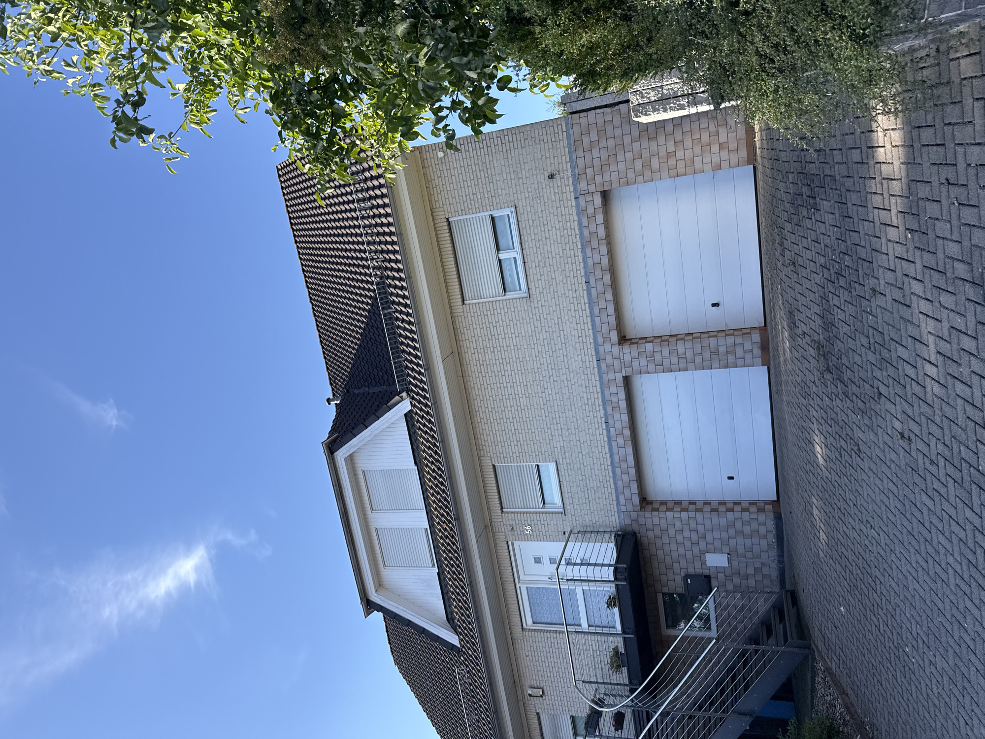 The image shows the exterior of a two-story house with a garage on the ground floor, located at Number 56 in Bad Sachsa, Germany. The house has a light-colored brick facade, a dark tiled roof, and a dormer window on the upper level. There are two white garage doors facing the viewer, and a metal staircase leading up to a balcony with a glass railing on the left side of the house. The driveway is paved with interlocking stones. A large green tree is visible on the right side of the image. The sky is clear and blue with a few wispy clouds.
