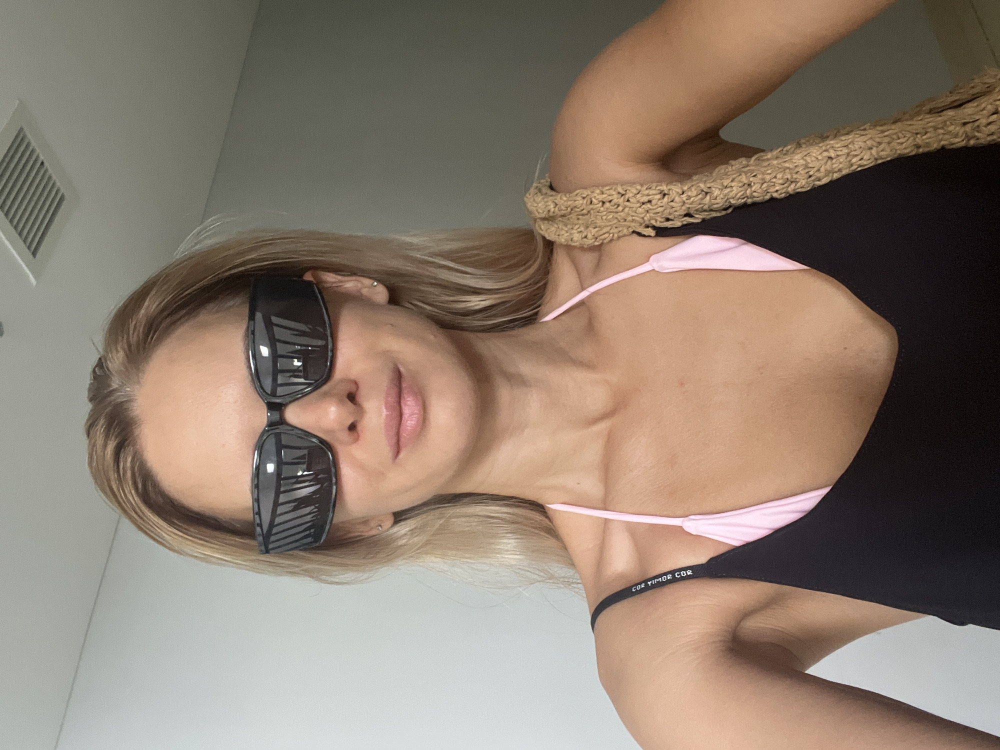 A fair-skinned woman with blonde hair pulled back and wearing large, dark, rectangular sunglasses stands facing the camera. She is wearing a black tank top over a pink bikini top, with a textured, woven strap draped over her right shoulder. The background is a plain, lightly colored wall with a ventilation grate visible on the ceiling. The lighting is bright and diffused, suggesting an indoor setting.