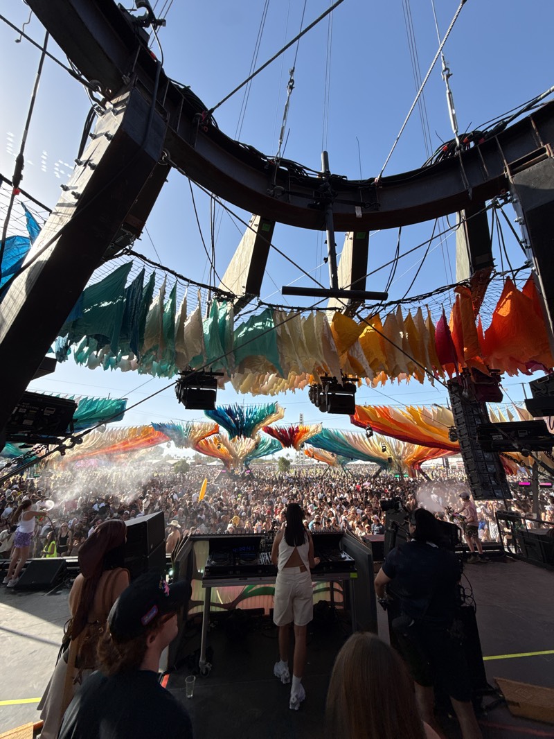The image captures a music festival, most likely Coachella, held in Indio, California. A DJ is visible on stage, facing a large crowd. Above the DJ, there is a structure with colorful decorations. There are many people in the audience. The sky is blue.