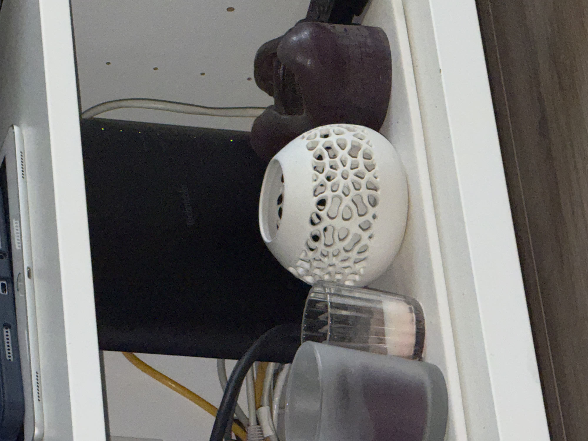 The image shows a close-up, eye-level view of a white shelf containing several objects. On the left, a dark gray glass tumbler holds a dark purple object, possibly a candle. Next to it is a clear glass votive holder with a pink candle. To the right of the votive holder is a white ceramic decorative object with an intricate, organic pattern of cutouts. To the right of the white ceramic object is a dark purple, irregular shaped object, possibly another candle or a decorative piece. In the background, a black electronic device with the word "technicolor" is visible, along with yellow and gray cables. The shelf is white and appears to be made of laminate or painted wood. The wall behind the shelf is light gray and has several small dark dots. The overall environment suggests an indoor setting, possibly a home or office. The image is cropped and does not include any views of Vilnius, Lithuania.