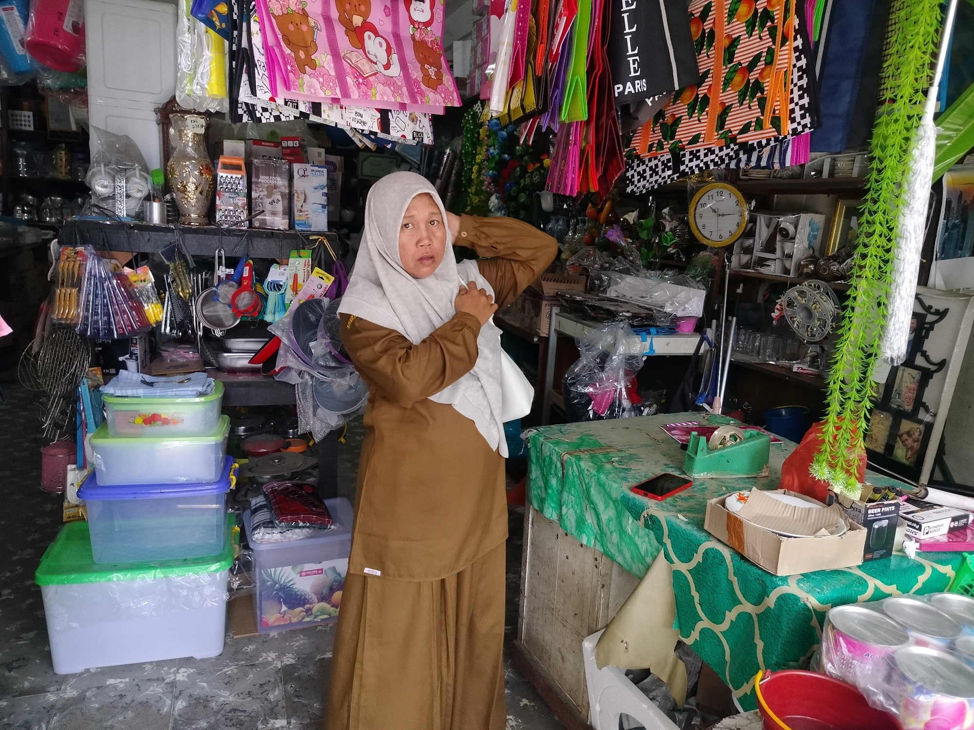 A woman wearing a light-colored hijab and a brown dress stands in front of a table covered with a green and white patterned cloth, in what appears to be a small shop in Perapat Hilir, Indonesia. The shop is filled with various items for sale. In the foreground, there are stacks of plastic storage containers. Behind the woman, the shop is cluttered with shelves holding vases, kitchenware, decorative items, and an assortment of packaged goods. Colorful fabrics and bags hang from the ceiling. A yellow wall clock is visible on a shelf. The overall environment suggests a local market or small retail establishment.