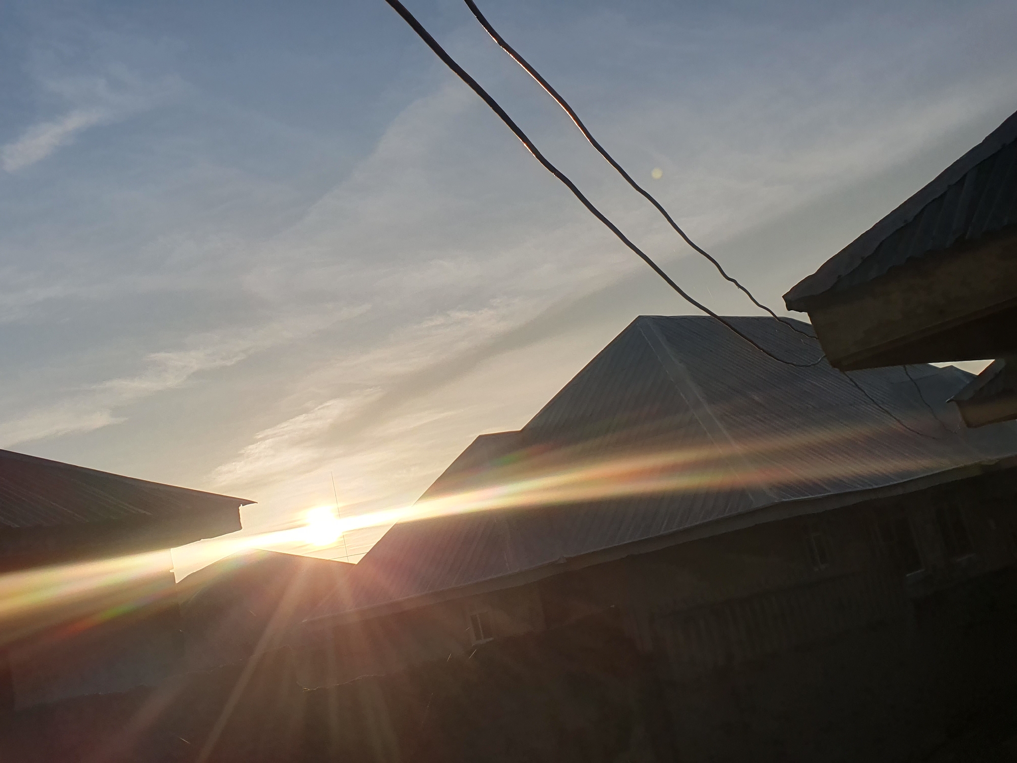 The image depicts a low-angle shot of rooftops against a bright, hazy sky, likely during sunrise or sunset. The sun is visible as a brilliant orb, casting strong lens flares and light streaks across the scene. Two parallel electrical wires traverse the upper portion of the frame, adding a man-made element to the natural sky and built environment. The rooftops appear to be constructed from corrugated metal, suggesting a functional rather than decorative purpose, and are characteristic of informal settlements or residential areas. The architecture, particularly the steeply pitched roofs and exposed construction elements, aligns with typical housing styles found in regions like Gura Top, Nigeria. There are no people visible in the image, and the focus is on the interplay of light and shadow on the structures and the sky. The overall impression is one of a developing urban or semi-urban landscape bathed in the warm light of the sun.
