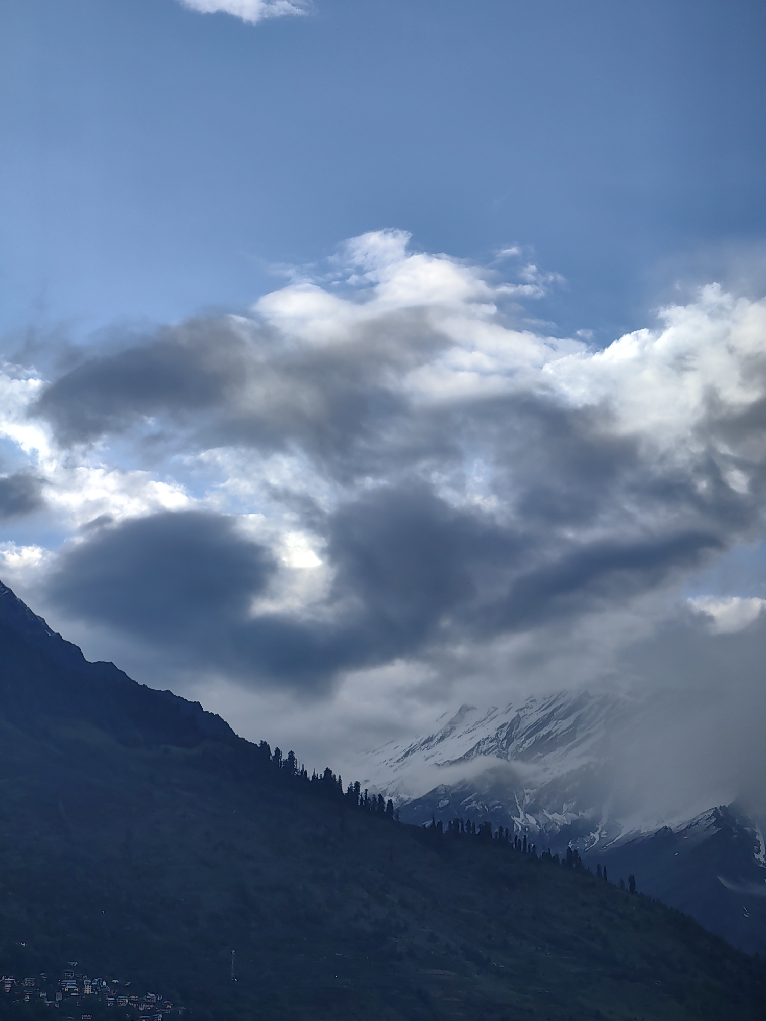A landscape image captures a view of mountains and clouds. The sky is a bright blue with scattered white and gray clouds. In the foreground, a dark, steep mountainside slopes upwards, dotted with evergreen trees along its ridge. At the base of the mountain, a cluster of small buildings, likely part of the town of Vashisht, India, is visible. In the background, snow-capped mountains rise, partially obscured by mist and clouds. The scene appears to be a natural vista with human settlement at the foot of the mountains.