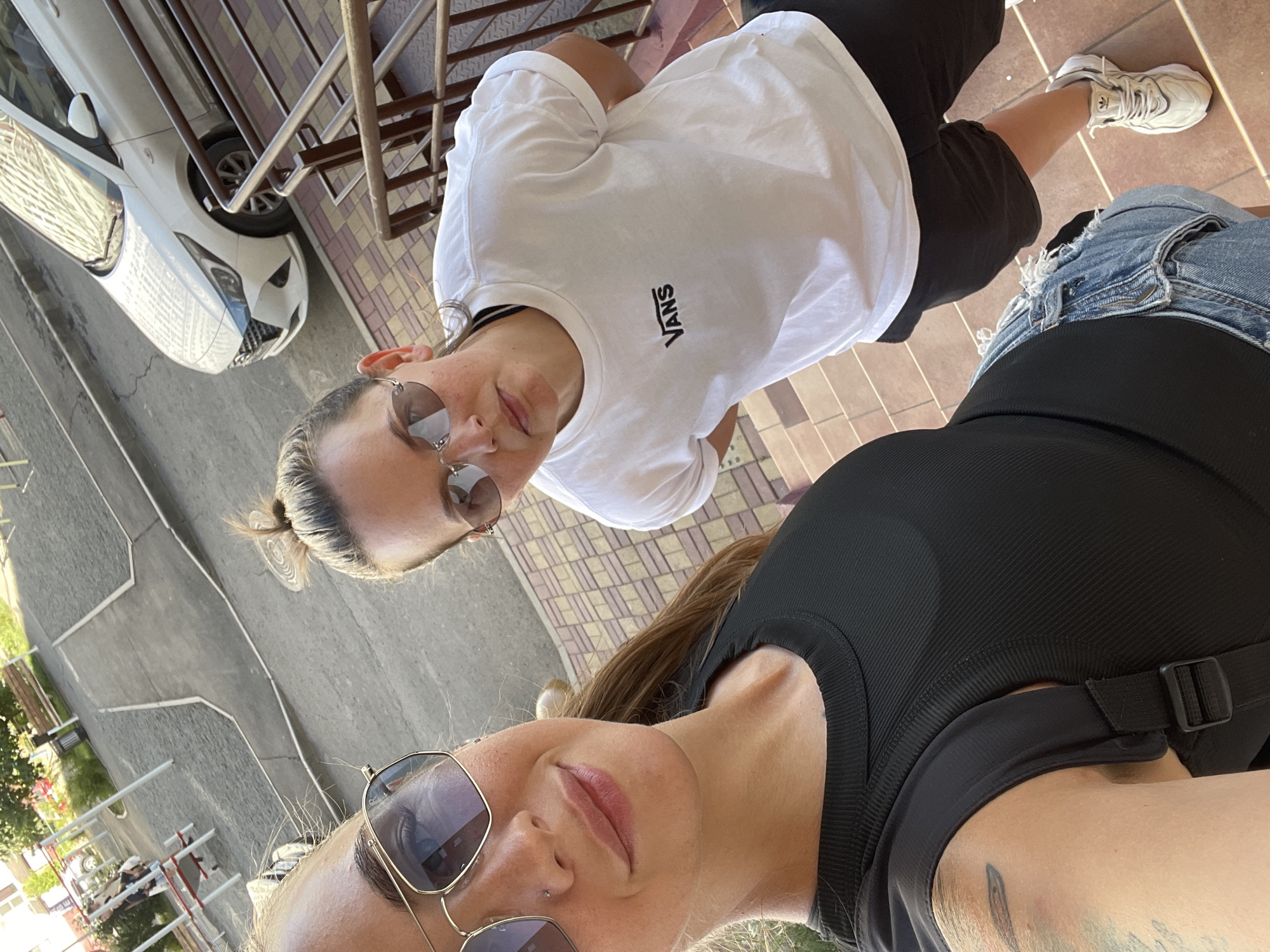 Two women, one in a black ribbed tank top and denim shorts, the other in a white Vans t-shirt and black shorts, pose for a selfie on an outdoor staircase in what appears to be a residential courtyard in Rostov-on-Don, Russia. The woman in the black top is in the foreground, wearing sunglasses and smiling. The woman in the white t-shirt is behind her, also wearing sunglasses, and looking towards the camera. A white car is visible parked on the street in the background, along with playground equipment and trees. The lighting suggests it is daytime.
