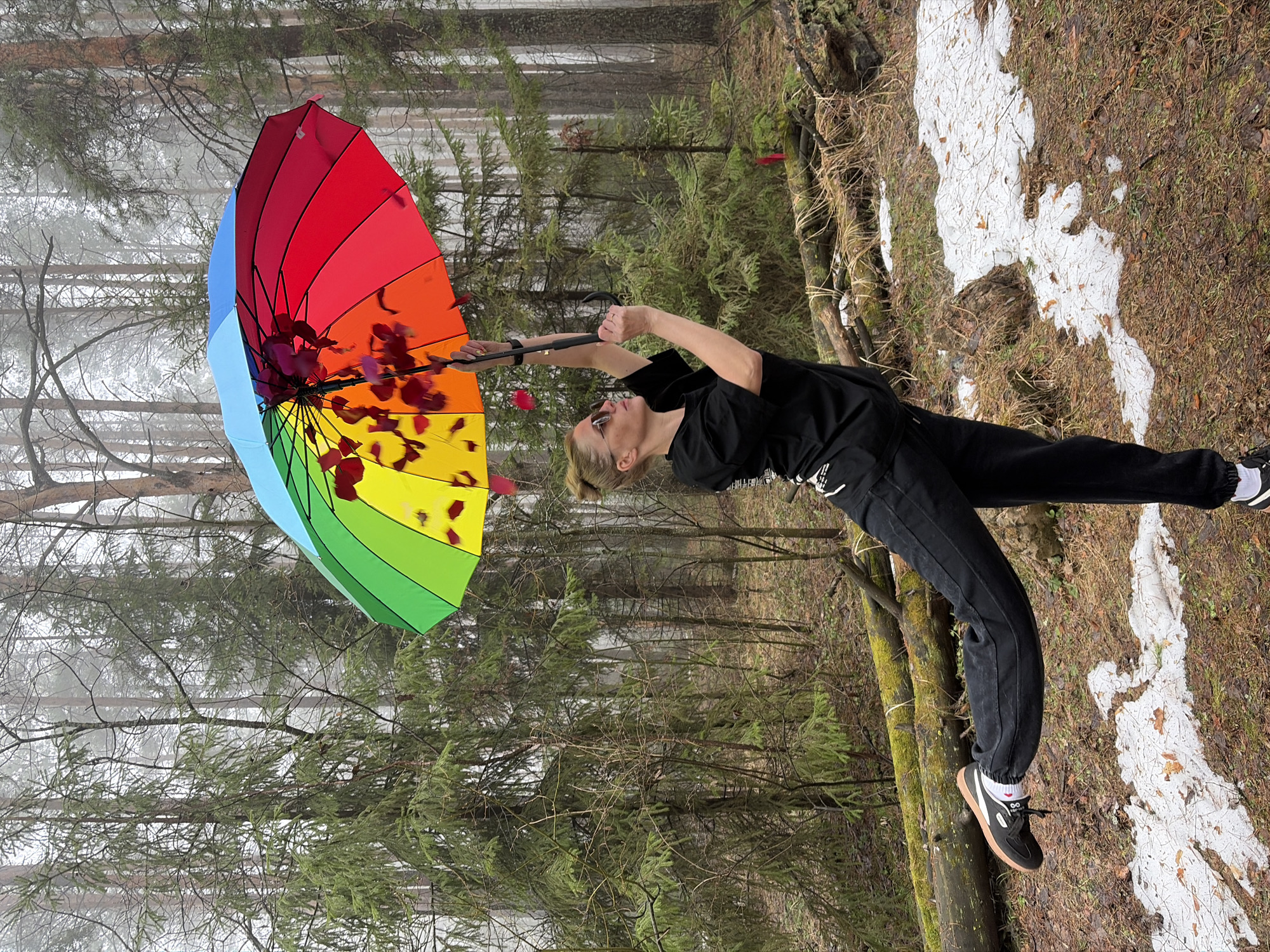 A woman in dark clothing is holding a multicolored umbrella in an outdoor setting. The background consists of trees and patches of snow on the ground. The location is Прикамский, Russia. The woman's body is arched, with her arms extended to hold the umbrella above her. The umbrella is filled with red rose petals. The scene appears to be set in a forest or wooded area.