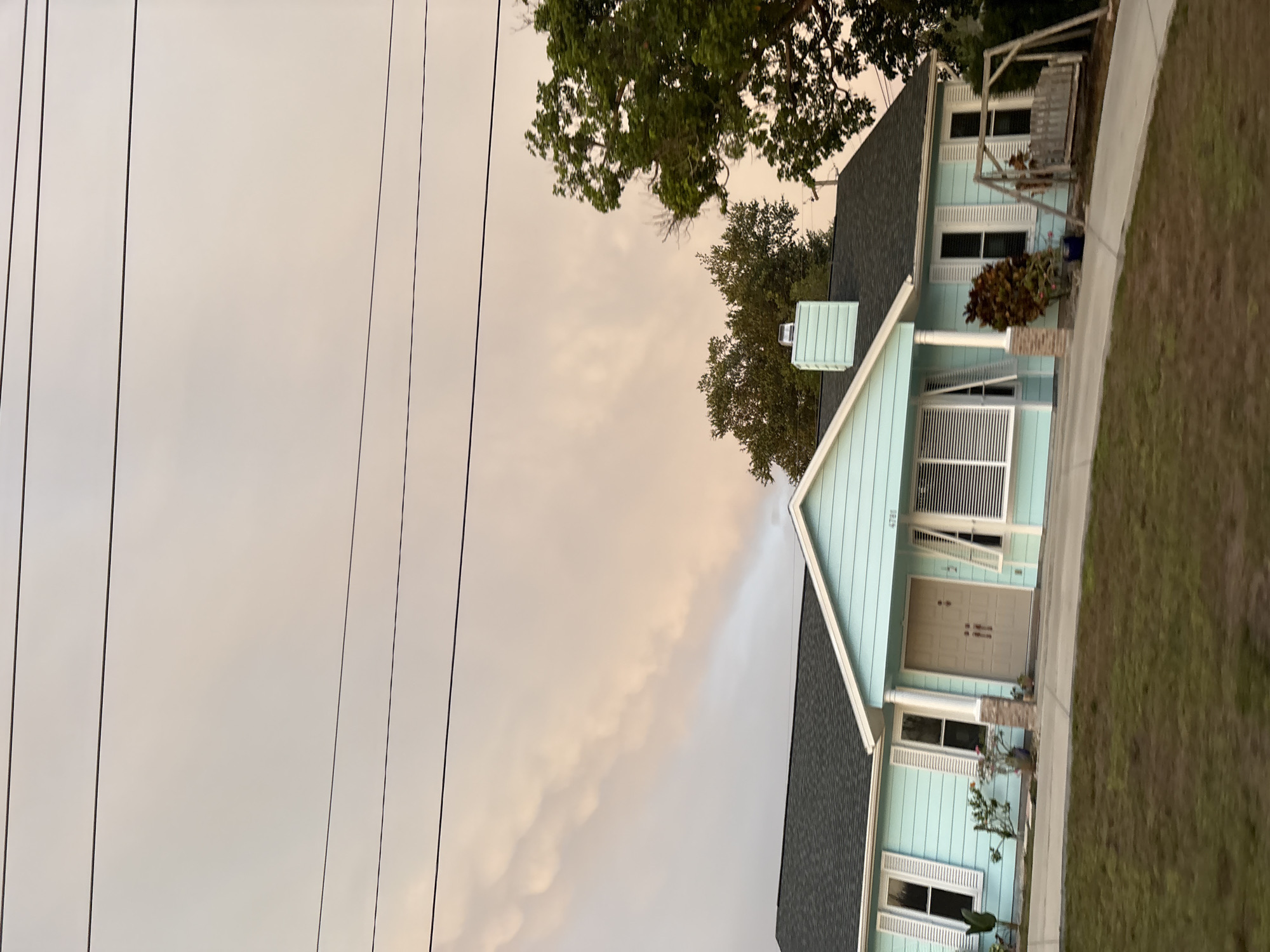 A light blue, single-story house with a dark gray roof and white trim is viewed from the front. The house has several windows with white shutters, a tan double front door, and a brick-accented support column. A chimney with light blue siding is visible on the roof. In front of the house, a driveway leads to the entrance. To the right of the house, there is a wooden porch swing. The yard in front of the house is covered with green grass. In the background, trees with green foliage are present. The sky above is filled with dramatic, light-colored clouds, possibly indicating a sunset or storm. Power lines are visible against the sky. The house is located at 4781.