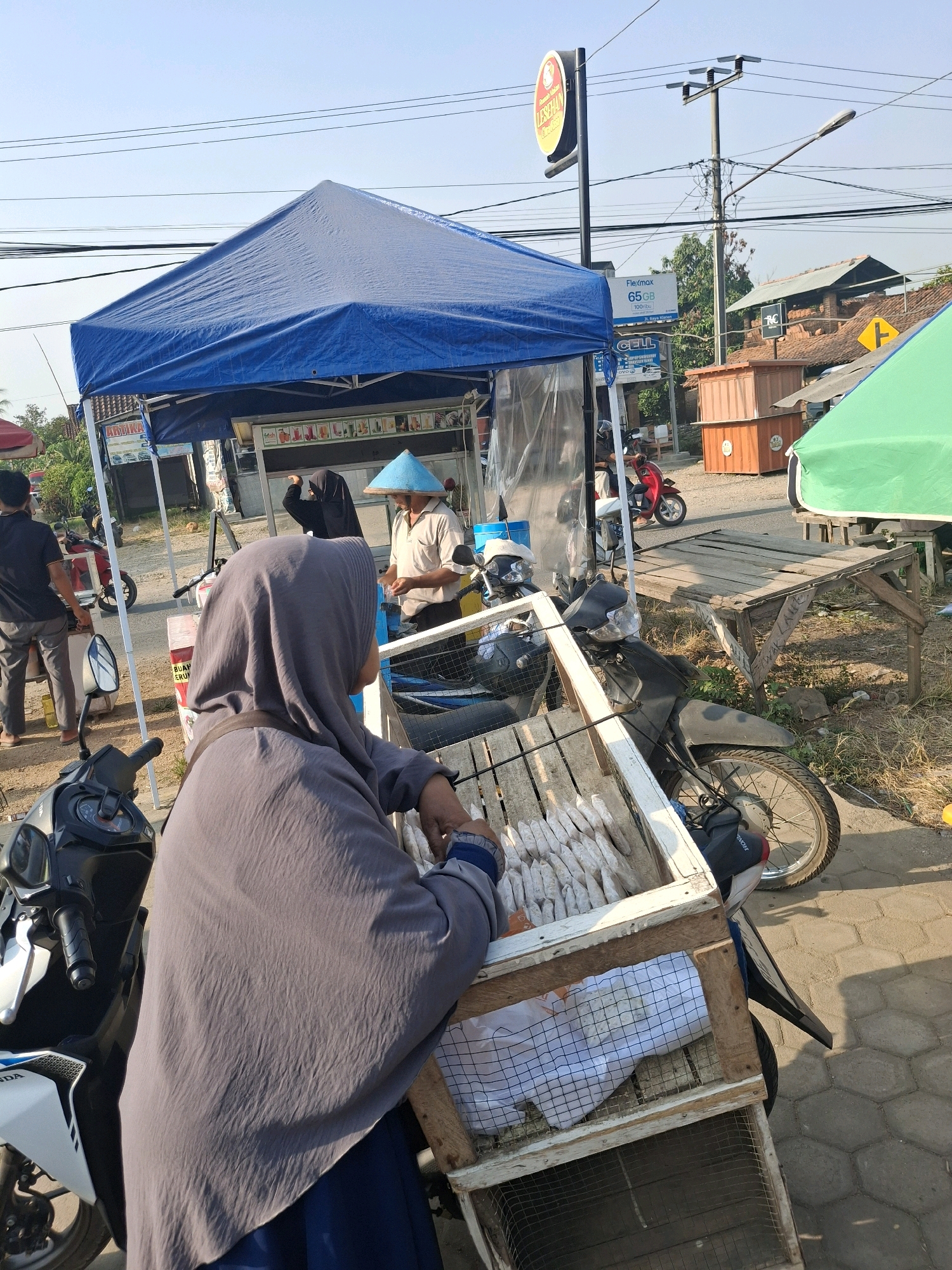 A woman wearing a grey hijab and a dark blue dress is seated in the foreground, facing away from the camera, looking at a stall. She is next to a motorbike. The stall has a blue canopy and displays food items. A man wearing a light-colored shirt and a conical hat is visible behind the counter, interacting with another person obscured by the woman. Several motorbikes are parked around the stall. In the background, there are buildings, electrical poles, and trees, indicative of a street scene in Bulurejo, Indonesia. The weather appears to be clear and sunny.