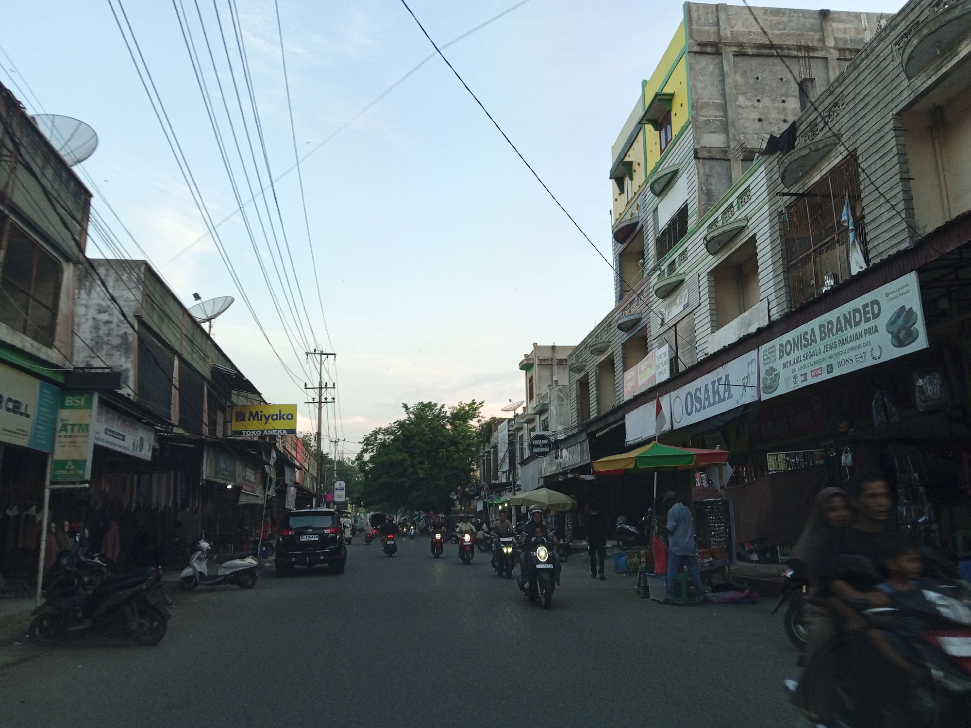This is a street-level photograph taken in Payakumbuh, Indonesia, during what appears to be late afternoon or early evening. The scene is bustling with traffic, primarily motorcycles.
On the left side of the street, there are several low-rise buildings housing shops. Signs visible include "BSI ATM" and "Miyako TOKO ANEKA." Motorcycles are parked along the sidewalk and on the road.
The right side of the street features taller, multi-story buildings. One prominent building has a yellow and green facade. Signs on the buildings include "OSAKA" and "BONISA BRANDED." A street vendor with a colorful umbrella is visible.
Several people are present, mostly riding motorcycles. A family is riding on one motorcycle in the foreground, with a man, a woman in a hijab, and two children. Other individuals are seen walking or standing near shops.
The road is paved and shows signs of wear. Overhead, numerous power lines crisscross the sky, contributing to a cluttered but characteristic urban landscape. Satellite dishes are mounted on some of the buildings. The sky is a pale blue with some wispy clouds.