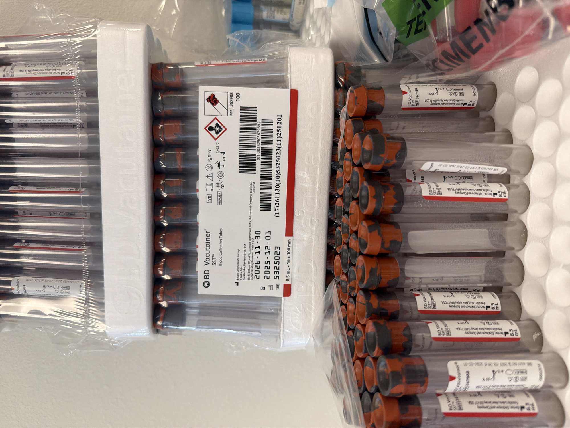 The image displays a close-up, eye-level, interior shot of medical laboratory equipment. Specifically, it features multiple rows of stacked BD Vacutainer SST blood collection tubes. The tubes are clear plastic with orange and black stoppers. A white label with black text is visible on the front of one of the upper rows of tubes, detailing product information such as "BD Vacutainer SST Blood Collection Tubes," expiration dates, lot numbers, and manufacturer information for Becton, Dickinson and Company. The tubes are housed in a white styrofoam-like packaging. The background is a plain, light-colored wall. In the lower part of the image, another layer of similar blood collection tubes is visible. To the right, partially obscured, are blue-capped vials and green and red labeled bags. The scene appears to be a controlled laboratory or storage environment. The photograph is taken in Beaverton, United States of America.