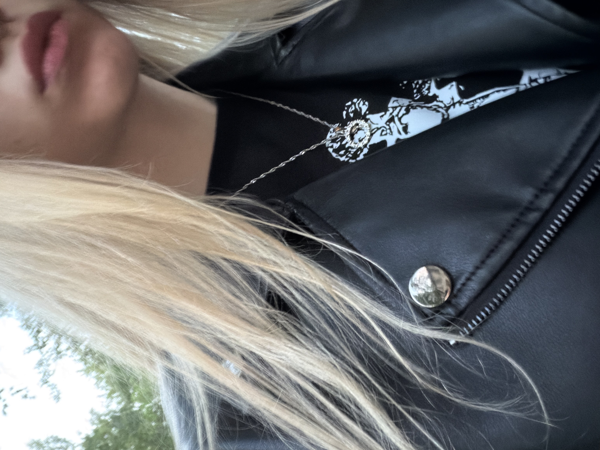 A partial view of a person wearing a black leather jacket and a necklace is captured. The individual has blond hair that partially obscures the jacket. The button of the jacket is visible, and a glimpse of a garment underneath with a white and black pattern can be seen. Only the lower part of the face is visible, showing only the lips. The exact location is not discernible from the given image.