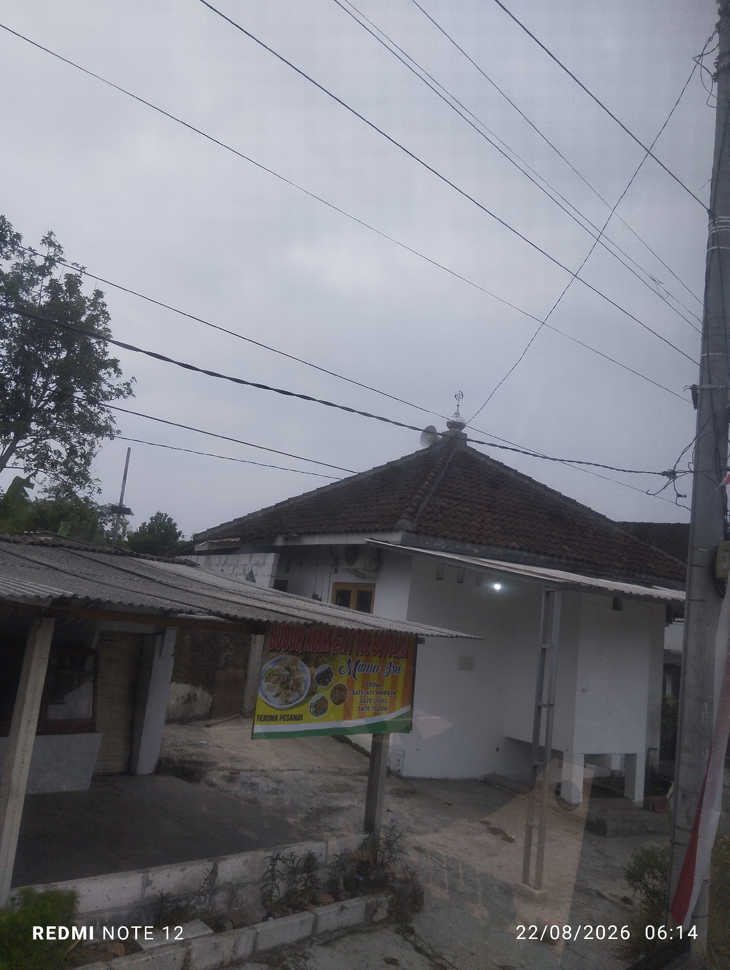 The image shows a cloudy sky with overhead power lines. In the foreground, there is a small, weathered building with a corrugated metal roof. To the right of this building is a larger, white building with a tiled roof, and a small mosque-like structure on its roof. A sign is visible on the left building, advertising "BUBUR AYAM EXIT TOLO Mama Isa," and listing menu items such as "SATE ATI AMPELA," "SATE USUS," and "SATE TELUR." The date and time "22/08/2026 06:14" are superimposed on the lower right of the image, along with the text "REDMI NOTE 12" on the lower left. The scene appears to be a roadside eatery or small shop in a rural or semi-rural area. The location is identified as Dipan, Indonesia.