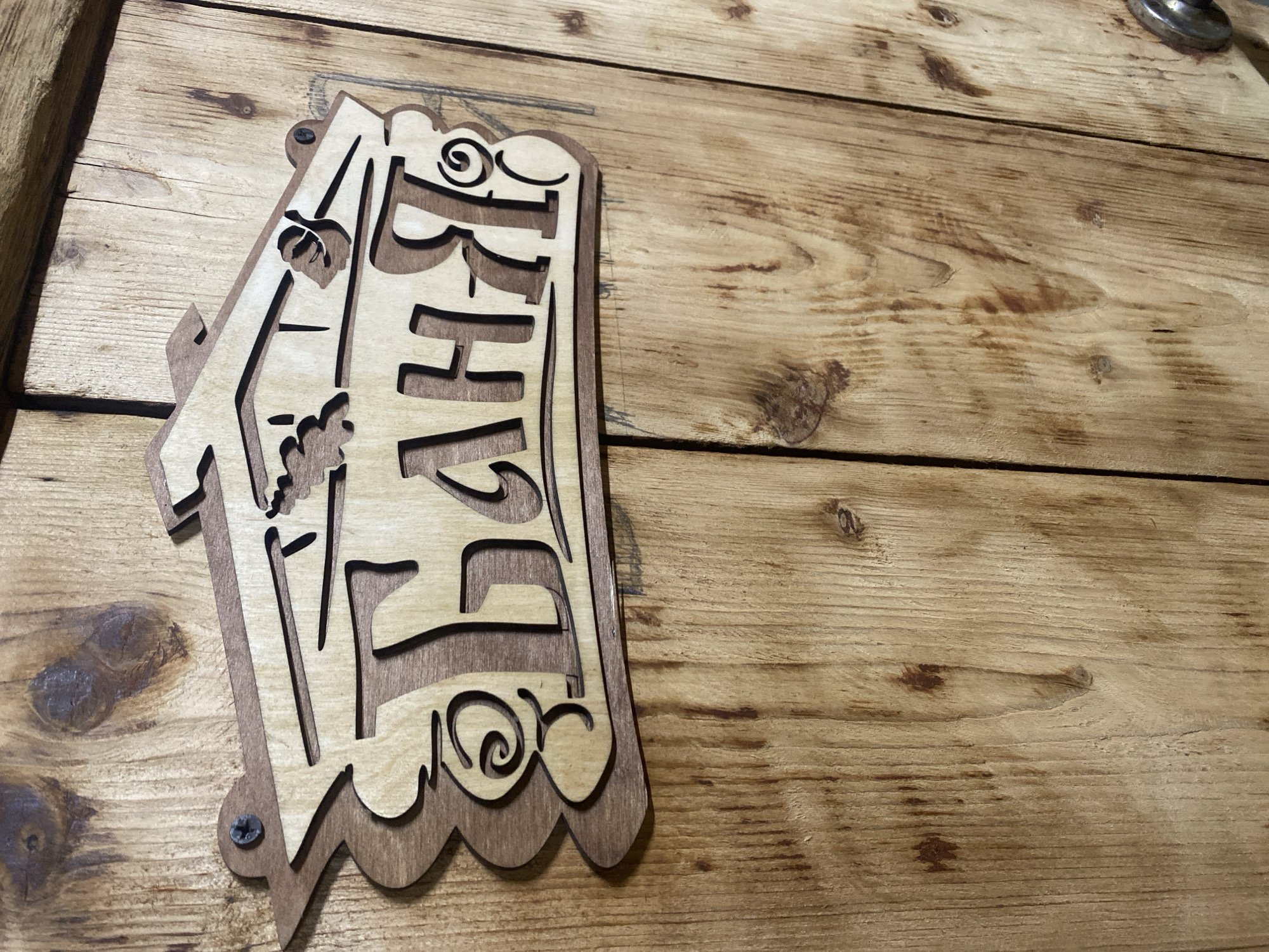 A close-up, eye-level shot captures a wooden sign hanging on a wooden wall. The sign, made of light-colored wood with a darker wood border, features carved Cyrillic letters that spell "БАНЯ" (Banya), meaning "bathhouse" in Russian. Decorative elements resembling leaves and swirls adorn the sign, which is attached to the wall with two screws. The wooden wall behind the sign is made of vertical planks, exhibiting a rustic texture with visible grain and knots, and is illuminated by natural light, casting soft shadows. The location is identified as Kisliakovskaya, Russia.