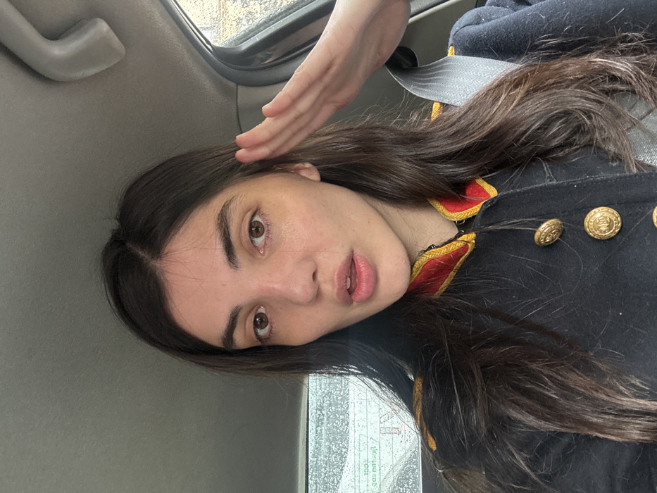 A female individual, appearing to be an adult, is seated in the passenger compartment of a vehicle. Her dark brown hair is long and parted centrally. She wears a dark, uniform-style coat featuring prominent red and gold trim on the collar and shoulders, with multiple brass-colored buttons, each bearing an emblem. Her right hand is raised to her forehead in a saluting gesture. A light gray seatbelt is fastened across her chest and right shoulder. The vehicle's interior features a gray headliner and a grab handle located above the right-side passenger window. Through the rain-streaked rear window, partial text "100% natural" and "gaz naturel" is visible. The external environment appears overcast. The presence of French text ("gaz naturel") indicates the location as Paris, France.