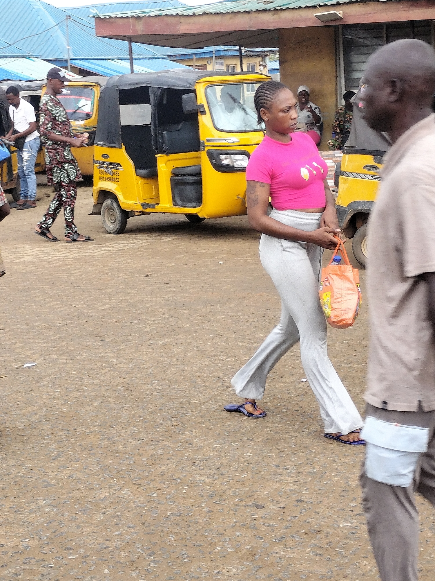 The image shows a street scene in Ikorodu, Nigeria. A woman in a pink crop top and gray pants walks across the street, carrying an orange plastic bag with a blue bottle inside. She has braided hair and a tattoo on her upper arm. In the background are several yellow "keke" or auto-rickshaws, a common form of transportation. Two men are walking in the opposite direction on the right side of the frame. Several other people are visible near the kekes in the background. The road surface appears to be paved and slightly uneven. Buildings are visible in the background, including a structure with a blue metal roof.