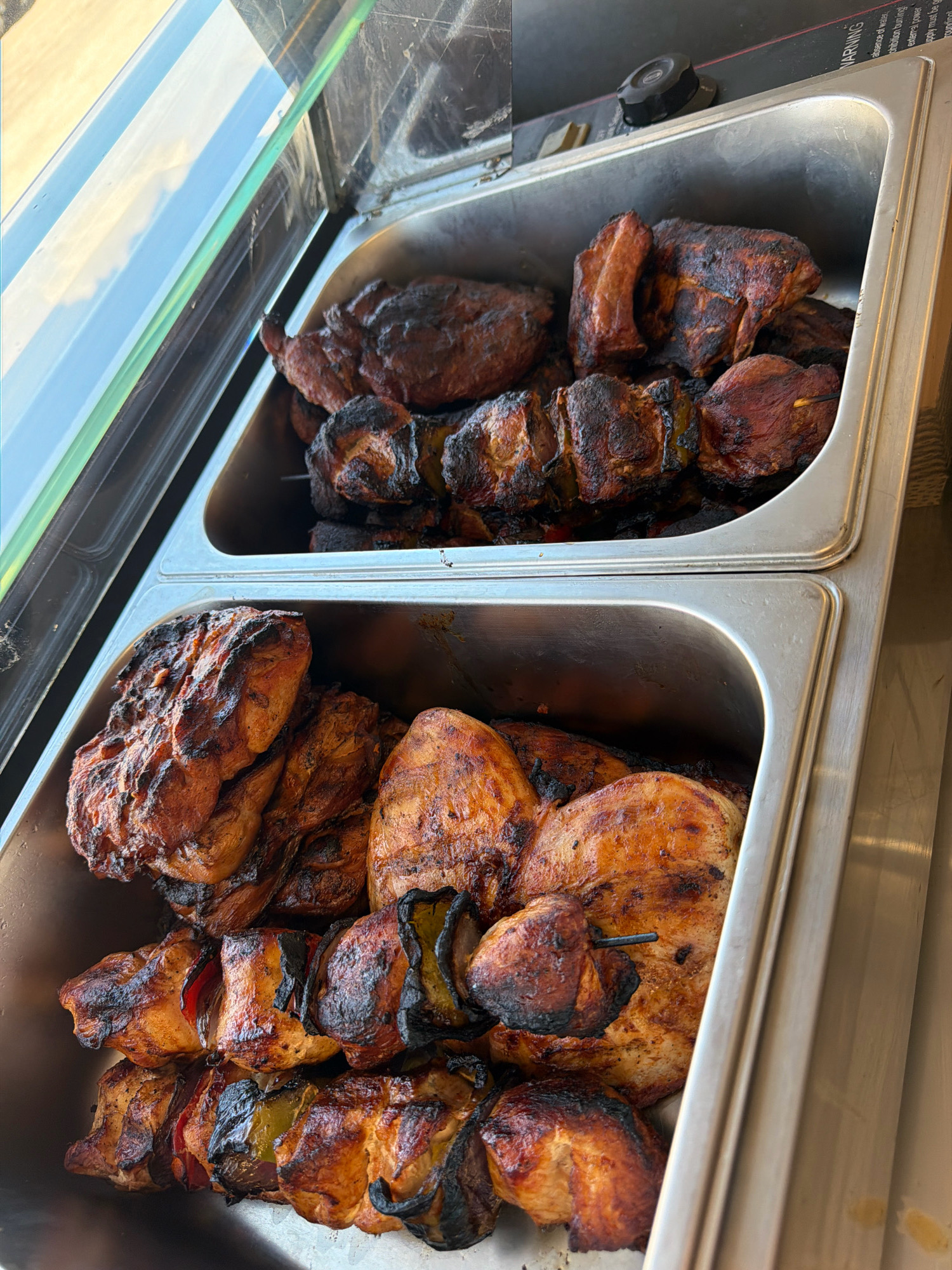 This is a close-up, eye-level, interior shot of two stainless steel food service trays filled with grilled meat. The top tray contains chunks of grilled meat, some on skewers, with visible charring. The bottom tray also contains grilled meat, including what appear to be chicken pieces and skewers with meat and vegetables. The food is well-lit and appears to be in a commercial setting. The background is out of focus, but suggests a modern interior with reflections on a glass surface in the upper left. There is a black dial visible on the right side of the top tray, suggesting a warming or cooking apparatus. The overall scene is clean and orderly, indicating a food preparation or service area. The food appears to be prepared and ready for consumption.