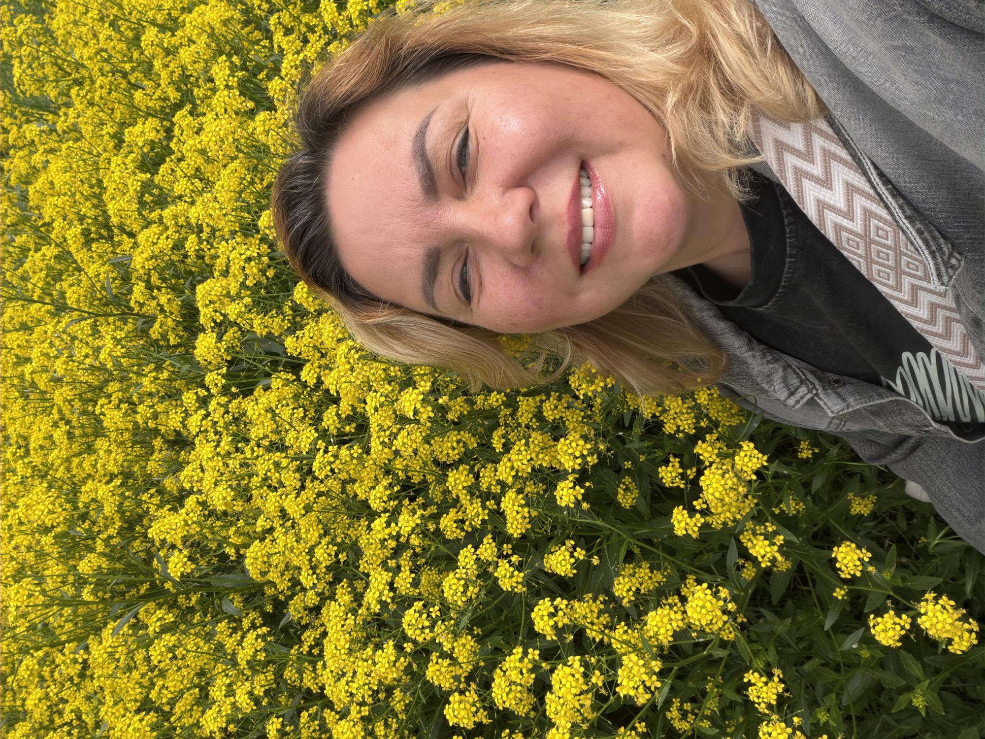 A middle-aged woman with blond hair is lying in a field of yellow flowers, possibly rapeseed. She is smiling at the camera. She is wearing a gray denim jacket over a black shirt with a light-colored design. A strap with a geometric pattern is visible on her jacket. The location is likely near Leningradskaya, Russia, given the prompt.