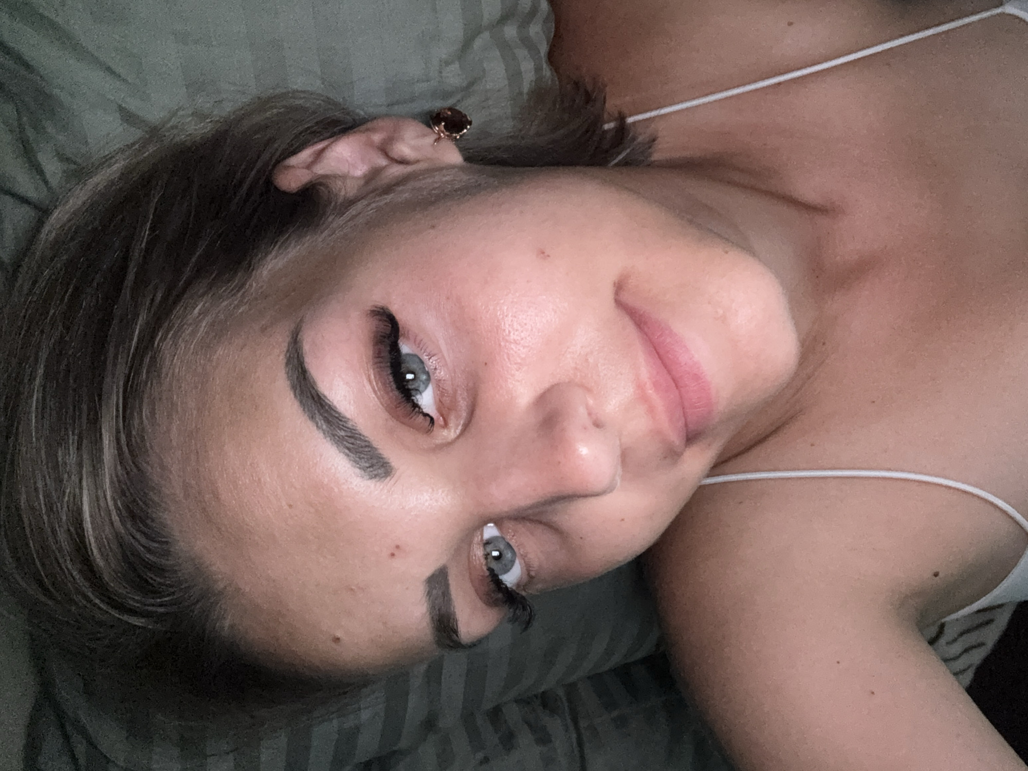 A close-up, high-angle selfie shows a young woman with fair skin and dark blonde hair lying down. She is wearing a white spaghetti strap top and a single, round, dark red stud earring in her right ear. Her left eye, a pale blue, is looking directly at the camera. Her eyebrows are dark and arched, and she has long, dark false eyelashes. Her lips are slightly parted, revealing a subtle smile. The background consists of a striped pillow in muted green tones, suggesting an indoor setting, possibly a bedroom. There are no other individuals or objects of note in the immediate vicinity. The image has a shallow depth of field, with the subject in sharp focus and the background slightly blurred. There is no visible connection to Yuzhno-Sakhalinsk, Russia.