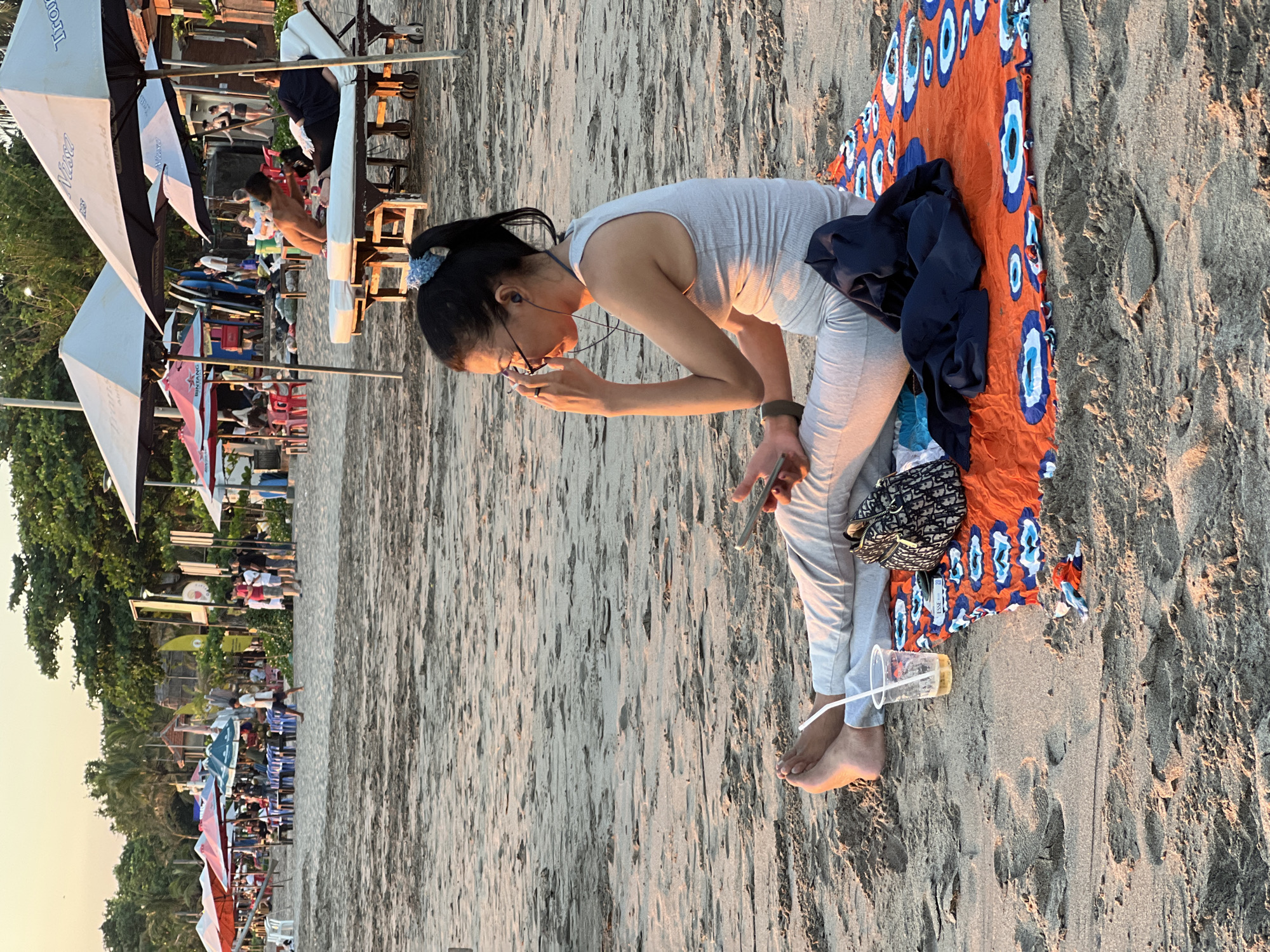 A beach scene in Kuta, Indonesia, is depicted during the golden hour, likely late afternoon or early evening. The image is rotated 90 degrees clockwise from its intended landscape orientation.
In the foreground, slightly right of center, a woman is sitting barefoot on a vibrant orange beach mat patterned with blue and white "evil eye" designs. She has dark hair tied in a ponytail with a light blue scrunchie and wears glasses, a light grey tank top, and matching pants. She is bending forward, engrossed with a mobile phone in her left hand, while her right hand is near her mouth. An earbud is visible in her right ear. Beside her on the mat are a dark blue jacket and a black and white patterned bag, possibly a Dior saddle bag. A clear plastic cup with a drink and a straw rests on the sand near her feet.
The background reveals a bustling beach setting. To the left, numerous beach umbrellas in white, red, and blue provide shade for people relaxing on chairs and at tables. Two prominent white umbrellas feature visible text, including "Bintang" and ".co.id," along with other partial Indonesian branding. Further back, rows of green trees line the beach, interspersed with beach shacks or kiosks and stacked beach equipment like surfboards. The sandy ground, illuminated by the warm, soft light, fills much of the frame, conveying a leisurely atmosphere at this popular tourist destination.
