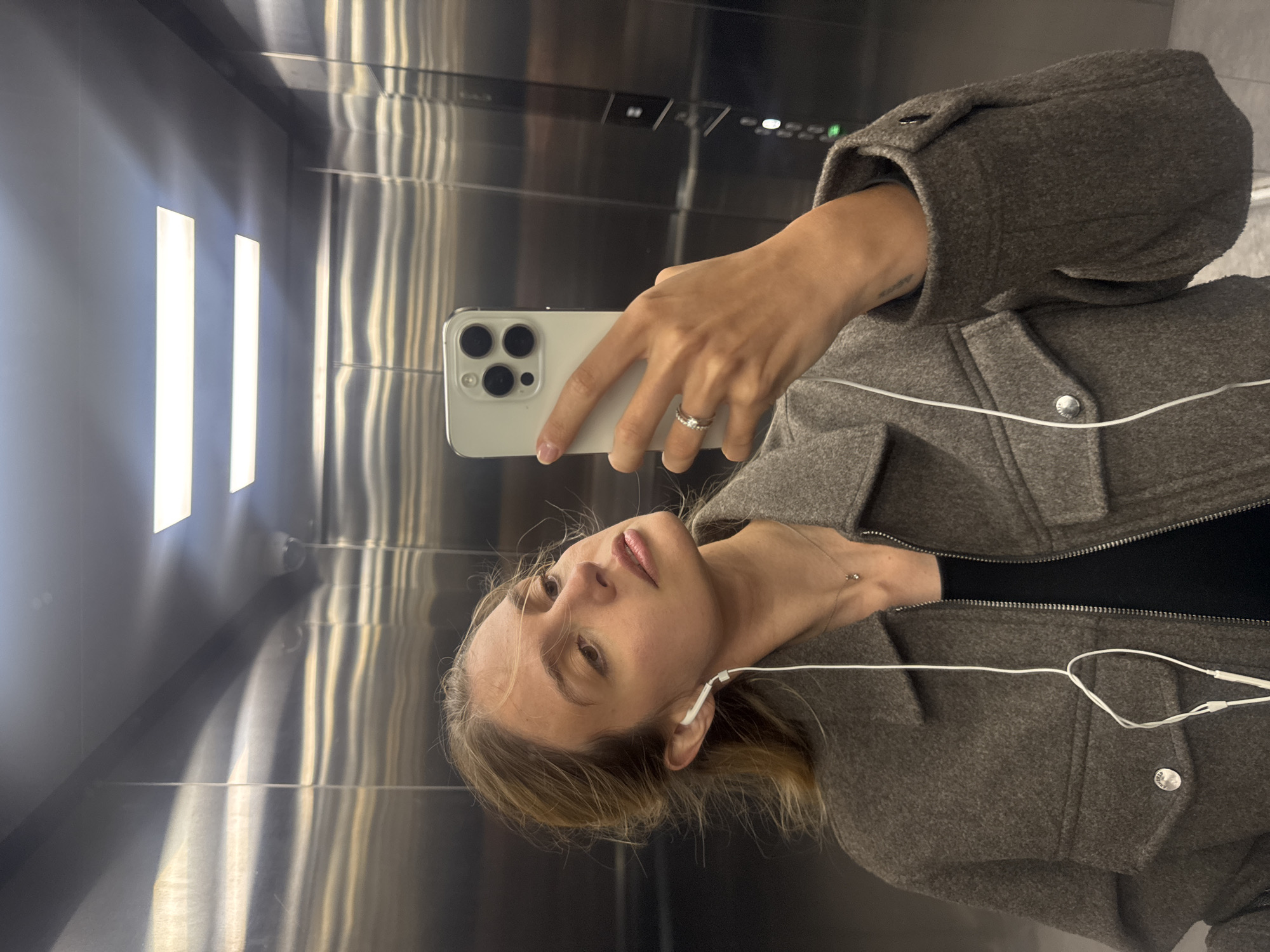 A young woman is captured in a selfie taken in a reflective surface, likely within an elevator. She appears on the right side of the frame, gazing towards the camera with a neutral expression. Her hair is light-colored and seems to be pulled back. She wears white wired earbuds in her ears.
In her right hand, she holds a light-colored smartphone, which features a triple camera array, characteristic of an iPhone. A silver-toned ring is visible on her left ring finger. She is dressed in a grey, textured jacket or coat over a dark top, with a delicate necklace and a small pendant around her neck.
The setting is an interior space with highly reflective, metallic walls, characteristic of an elevator, with bright, vertical strip lighting visible on the left side. These lights and other elements of the elevator interior, including what appears to be a control panel with buttons, are reflected across the polished surfaces. The overall lighting is bright and even. No discernible text is visible in the image.