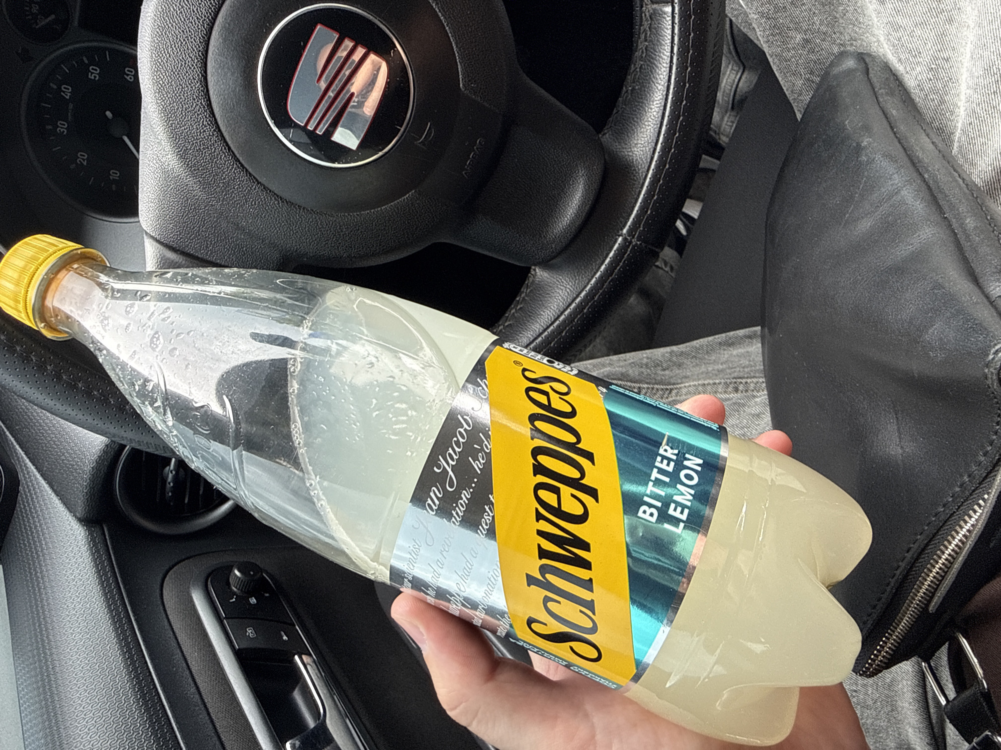 A close-up shot captured from inside a car in Donets, Ukraine, shows a person's right hand holding a partially consumed bottle of Schweppes Bitter Lemon. The bottle is clear plastic with a yellow cap, and its label prominently features the yellow "Schweppes" logo and "BITTER LEMON" in green. Visible text on the label also includes "An Jacob Schweppe had a revelation... he'd had a quest" related to carbonation.
The setting is the interior of a car, specifically a SEAT model, indicated by the logo on the steering wheel, which is partially visible in the upper left. The dashboard speedometer or tachometer shows markings from 10 to 60, with the needle near zero. A door panel with textured trim, an air vent, and window controls are visible on the left. The person's leg, clad in grey denim jeans, is visible on the right, next to a black leather bag or purse with a silver zipper. The lighting suggests it is daytime. No specific activity beyond holding the drink is discernible, nor are any emotions conveyed.