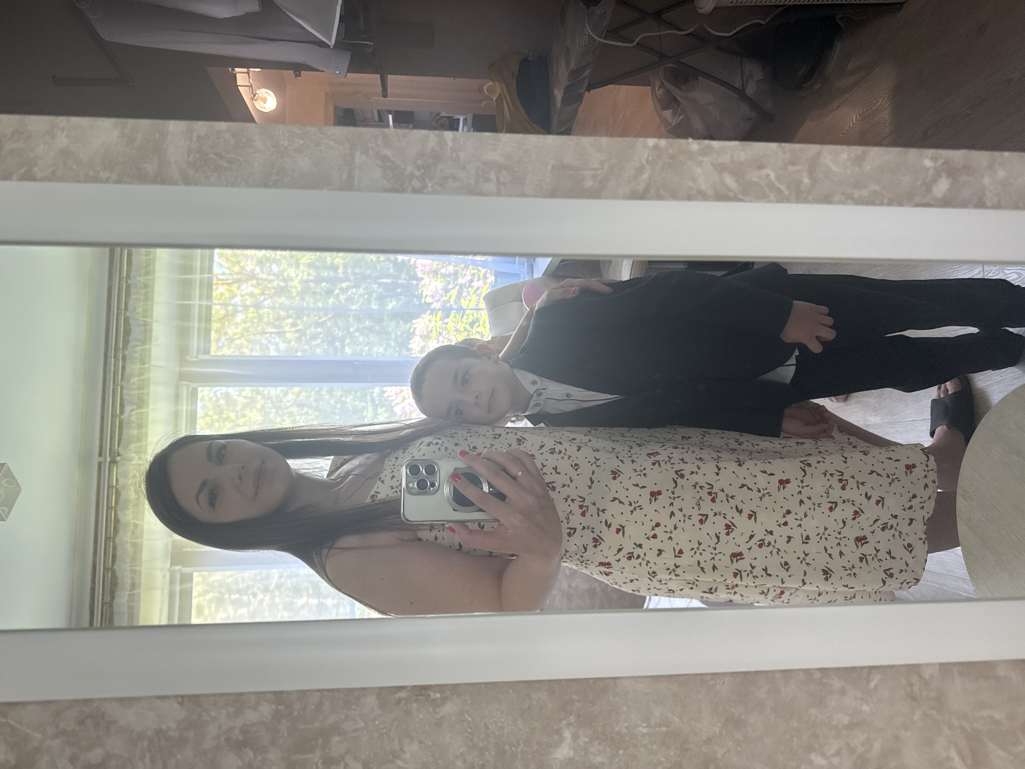 A woman is taking a selfie in a mirror with a young boy. The woman is wearing a white dress with a red floral pattern and holding a silver smartphone. The boy is wearing a black suit and a white shirt. The background shows a room with a window and a glimpse of the outdoors. The location may be Razdolie, Russia.