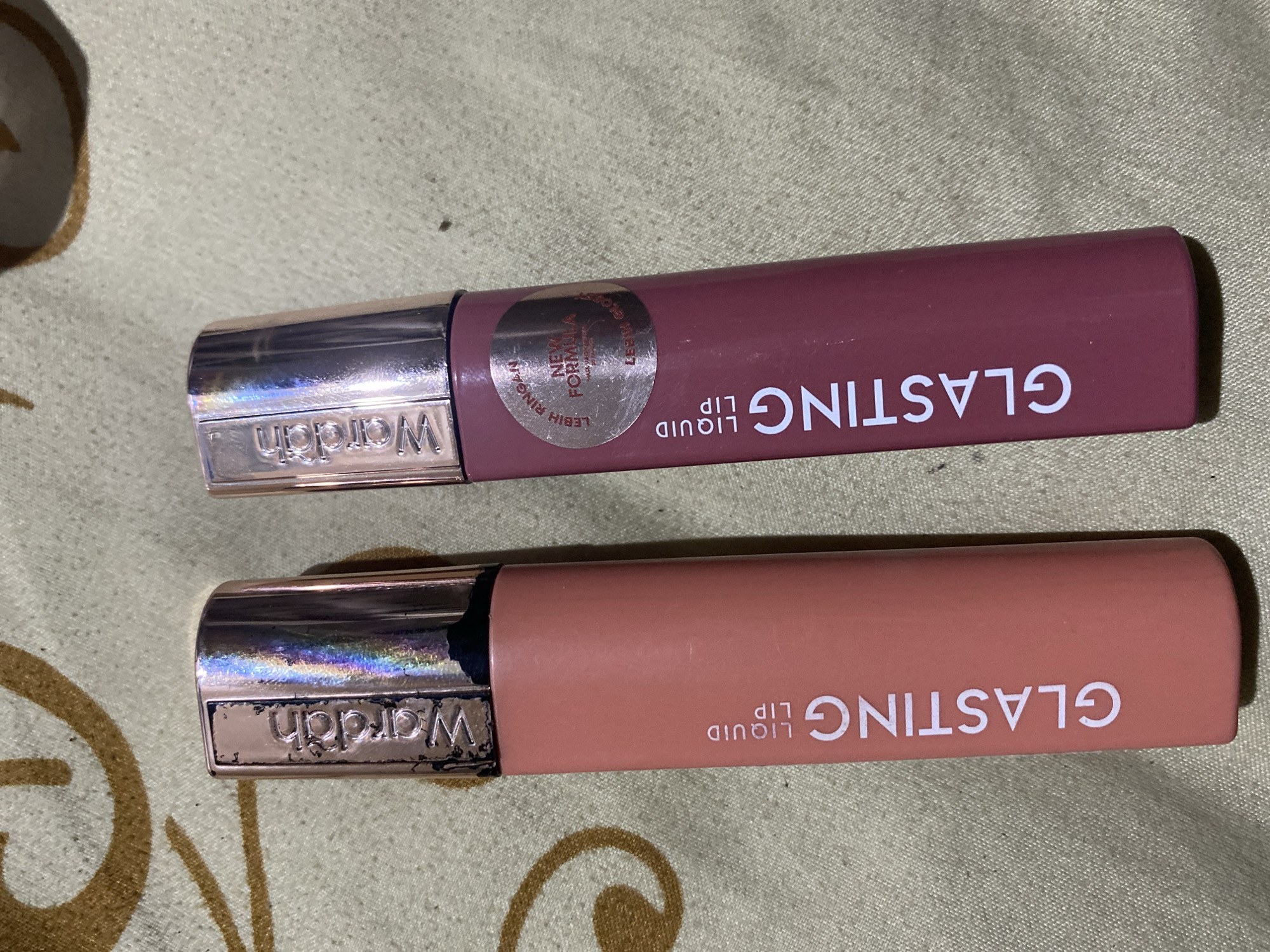 Two tubes of Wardah "Glasting Liquid Lip" are shown. The tube on the left is coral-colored and has the text "GLASTING LIQUID LIP" vertically printed on it. The tube on the right is a dusty rose color and also has "GLASTING LIQUID LIP" printed vertically. It also has a circular sticker that reads "LEBIH RINGAN NEW FORMULA LEBIH GLOSSY." Both tubes have a silver cap with the "Wardah" logo imprinted. The items are placed on a textured green fabric. The scene is likely indoors, in a domestic setting, and not specifically identifiable to Ta', Indonesia.