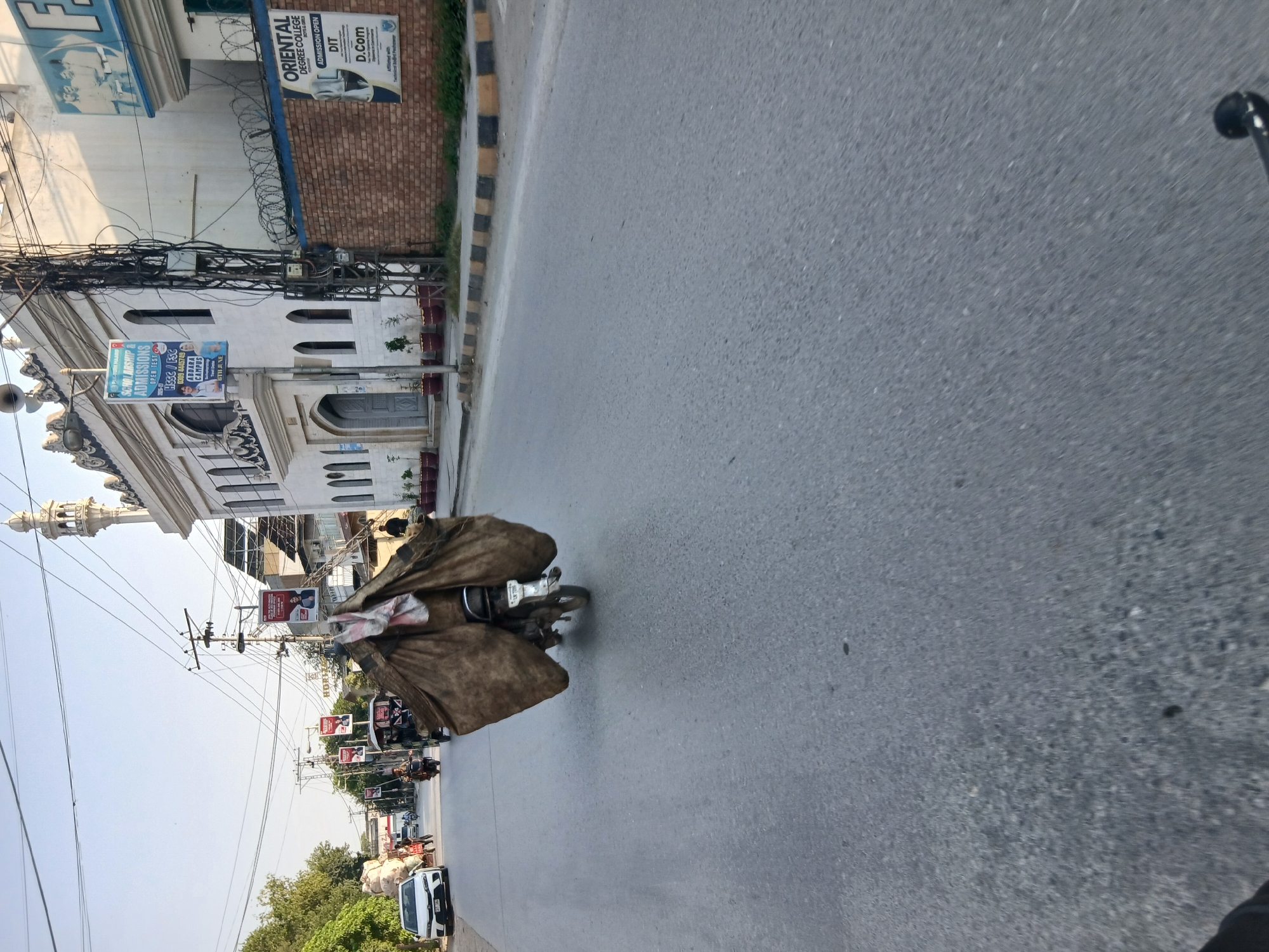 The image depicts a street scene in Peshawar City Tehsil, Pakistan. A motorcycle rider, with a large, brown, tarp-like covering extending outwards from the back of the motorcycle and obscuring the rider, is the central subject. The tarp appears to be carrying a significant load. The motorcycle is in motion on an asphalt road. On the right side of the road, a white building with a minaret and arched windows, likely a mosque or religious institution, is visible. Adjacent to this building is a brick wall topped with barbed wire. A sign for "Oriental Degree College" is affixed to the brick wall. Further down the street, other vehicles, including a white car and a rickshaw, are present. Utility poles with numerous wires are scattered across the scene. The environment appears to be an urban or semi-urban setting, with clear skies overhead.
