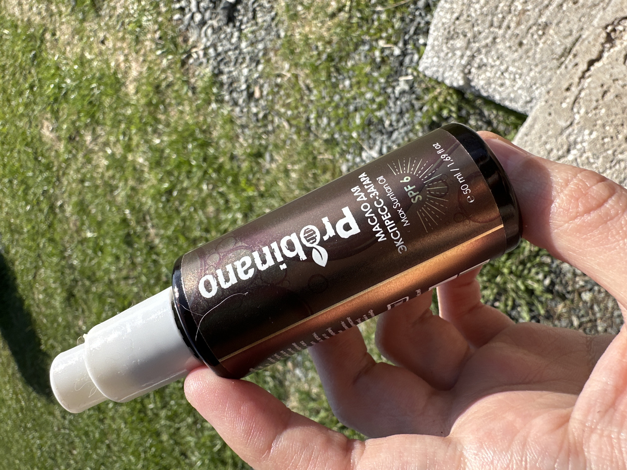A human hand holds a bottle of "Probinano" Max Suntan Oil SPF 6, which is a dark-colored bottle with a white pump dispenser. The text on the bottle is partially in Russian. The bottle is held against a background of green grass and a patch of light-colored gravel. The scene appears to be outdoors in daylight. There is no information available to conclude that the location is Kharnavy, Russia.