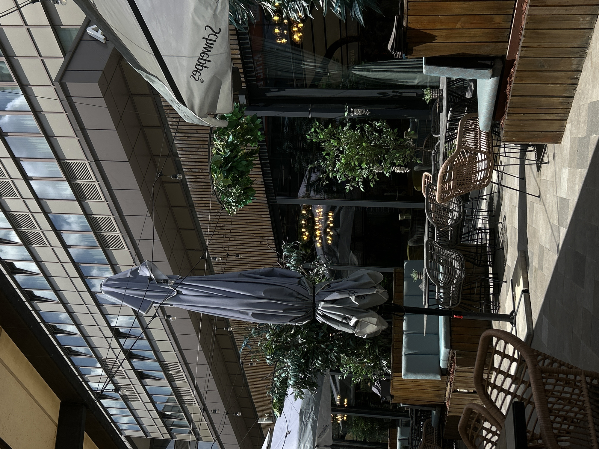 The image depicts an outdoor seating area of a café or restaurant in Sofia, Bulgaria. The location appears to be near a modern office building with a reflective glass facade, catching reflections of the sky. The seating area is shaded by large umbrellas. Several wicker chairs are arranged around tables. Various potted plants provide decoration. The area is paved with gray tiles. The time is likely during the day, as the light suggests. There are no people visible in the immediate scene.