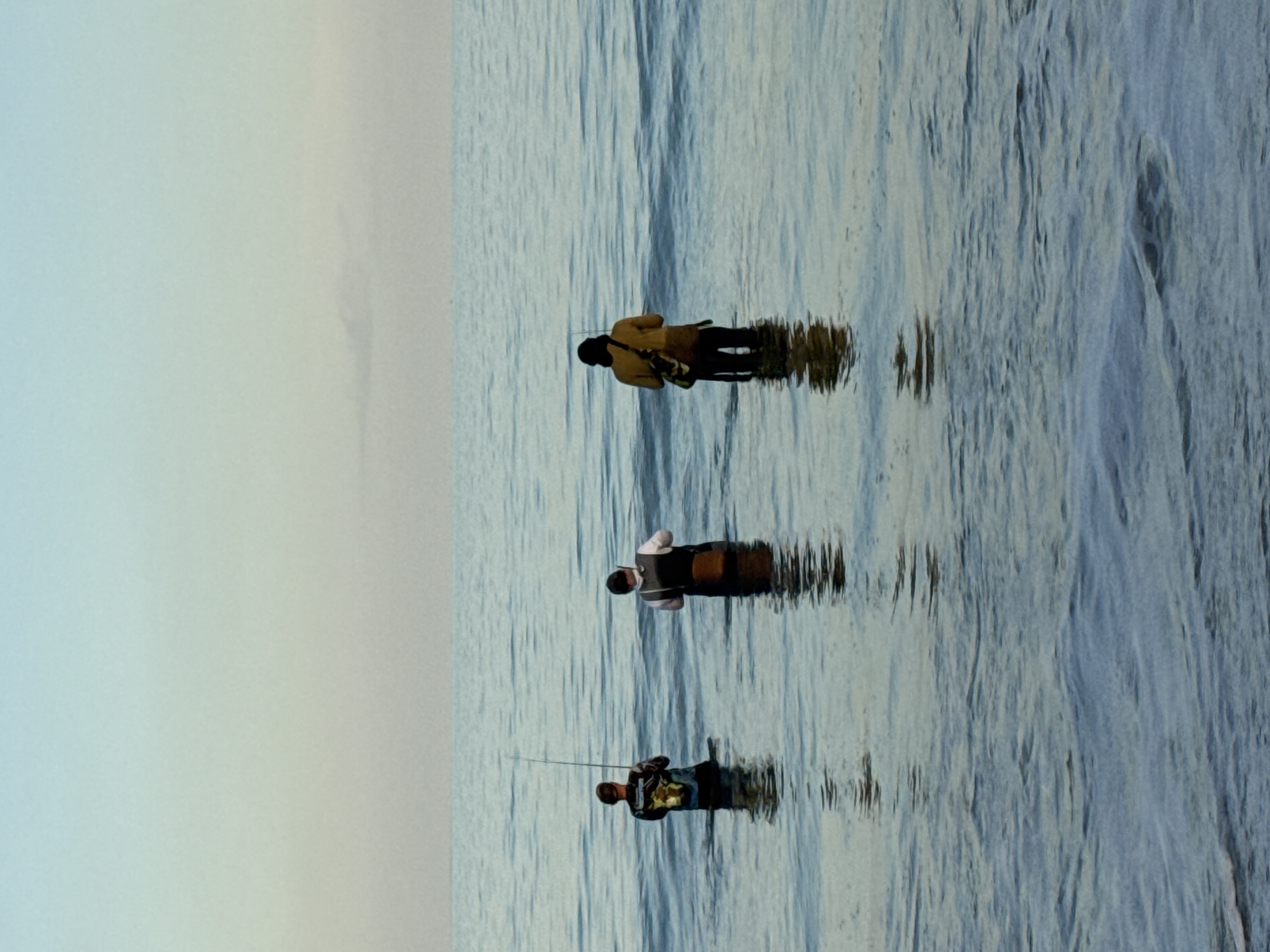 Three people stand in shallow water in the ocean. The leftmost person is holding a fishing rod. The person in the middle is wearing a dark vest over a white shirt. The person on the right is wearing a tan shirt and carrying a bag. The water is wavy. The sky is hazy. The location is likely Kuta Selatan, Indonesia, based on the image query.
