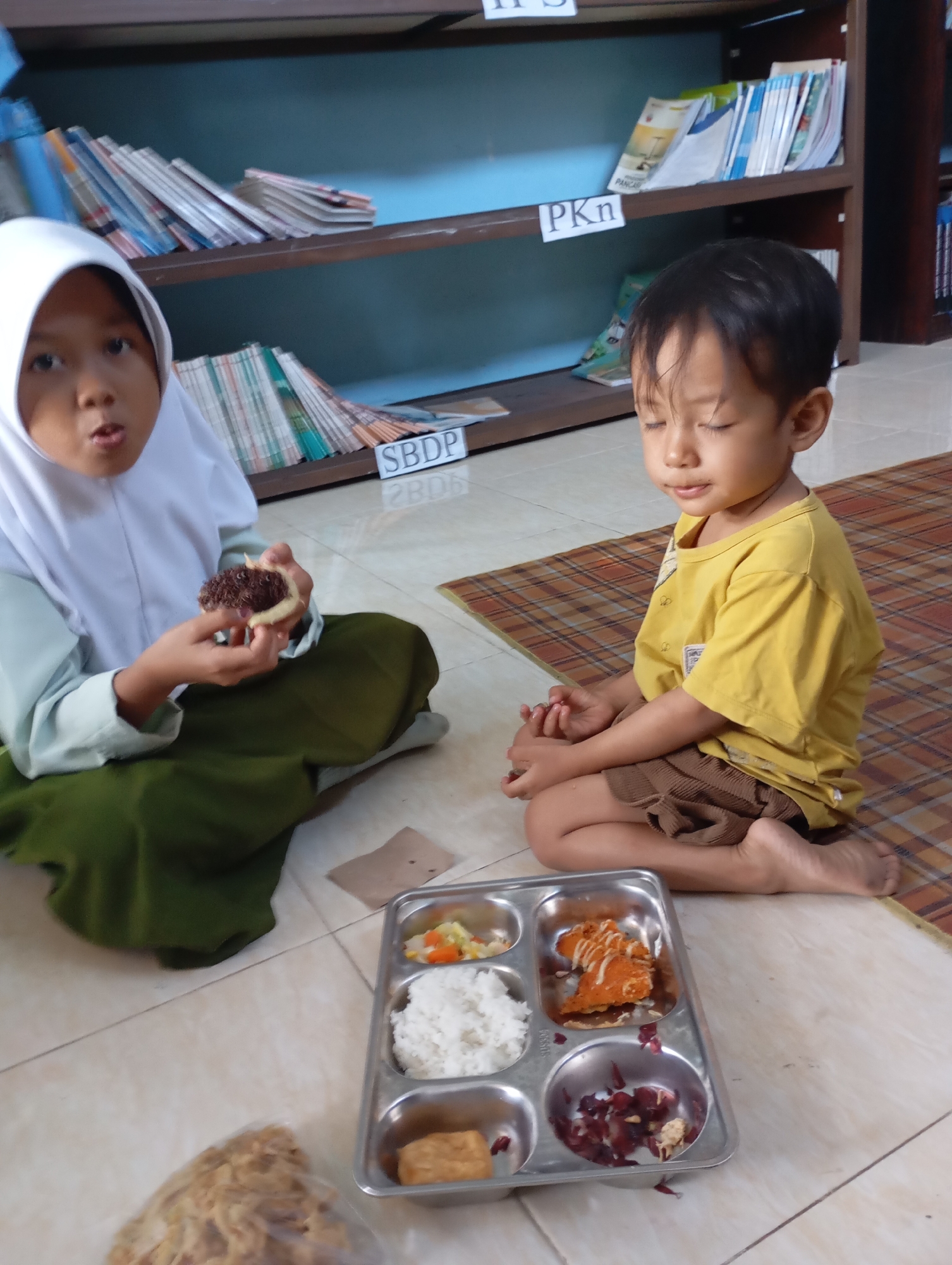 In a room with tiled flooring and bookshelves in the background, two young children are seated on the floor. The child on the left, wearing a white hijab and a green robe, is holding a donut-like pastry with chocolate sprinkles and appears to be eating it. The child on the right, wearing a yellow t-shirt and brown shorts, is sitting with their eyes closed, seemingly resting. In front of the children is a four-compartment metal tray containing rice, vegetables, a piece of fried food, and some red berries. The bookshelves contain numerous books and papers. The scene is likely an educational institution or a home environment. The presence of the children and the meal suggests a time of sustenance or a break period.