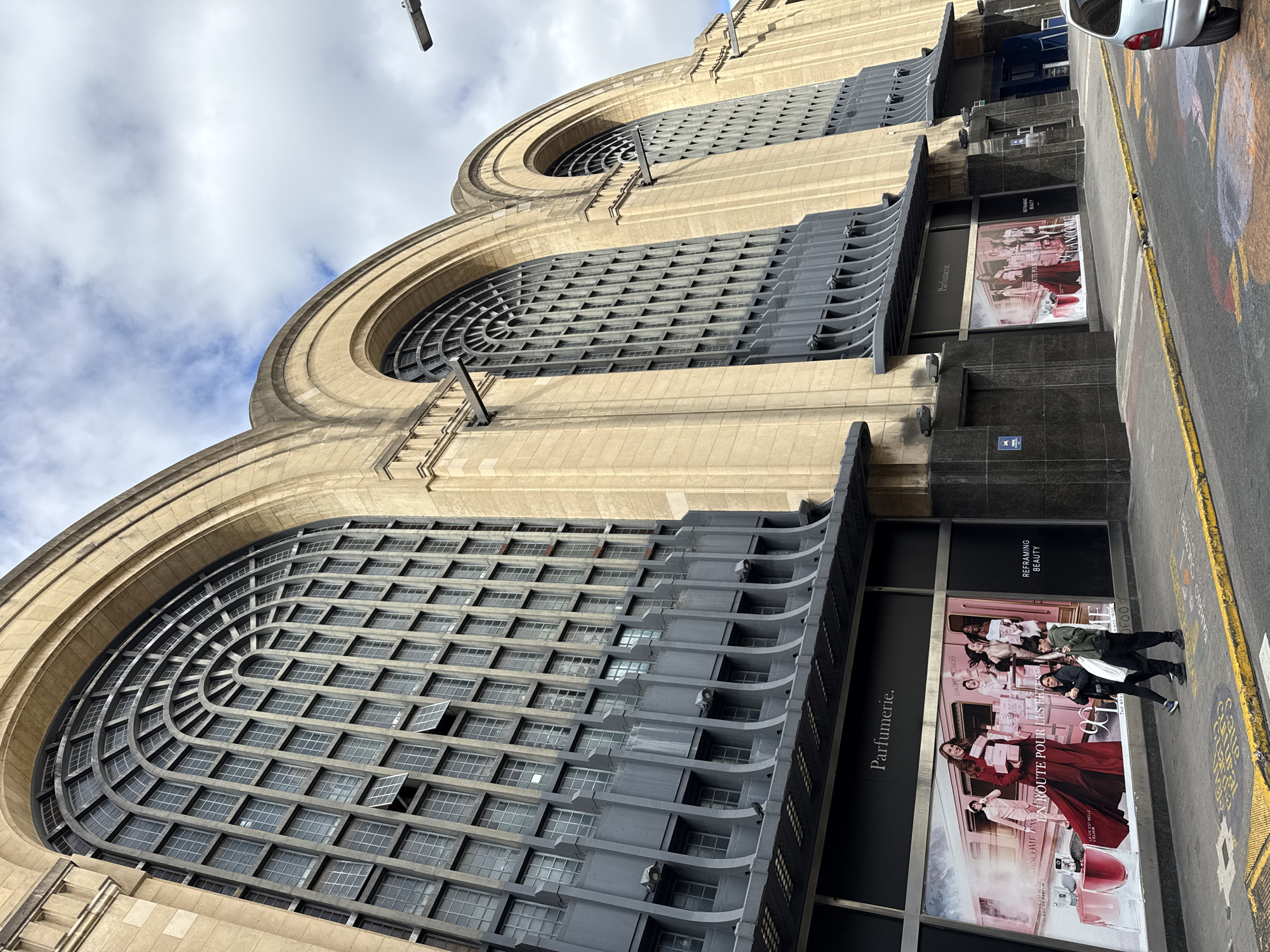 This media file presents a striking architectural facade from a low-angle perspective, likely on a street in Buenos Aires, Argentina. The prominent subject is a large, multi-story building featuring a distinctive Art Deco or Art Nouveau style, constructed from light-colored stone. Its most notable features are the grand, arching window sections, intricately designed with a grid of numerous square and rectangular panes, some of which are slightly open. These windows are recessed behind a series of grey, horizontal architectural elements that provide a layered, curved effect.
At street level, the building is clad in darker material, possibly granite or marble, housing large storefronts. These display advertisements for "Parfumerie" and the brand "LANCOME," with visible text including "LA VIE EST BELLE," "EN ROUTE POUR LES FÊTES," and "REFRAMING BEAUTY." Two individuals, an adult and a younger person, are standing on the sidewalk directly in front of one of these large ads.
The scene is captured during the daytime under a partly cloudy sky. In the foreground, a sidewalk with yellow painted lines and some faded markings, including partial words like "CALLE GRAL" (likely "Calle General") and "CALLE CONC" (possibly "Calle Corrientes"), suggests an urban street setting. A silver car is partially visible on the street to the right. The image highlights the detailed craftsmanship of the building's architecture against the backdrop of a vibrant city street.