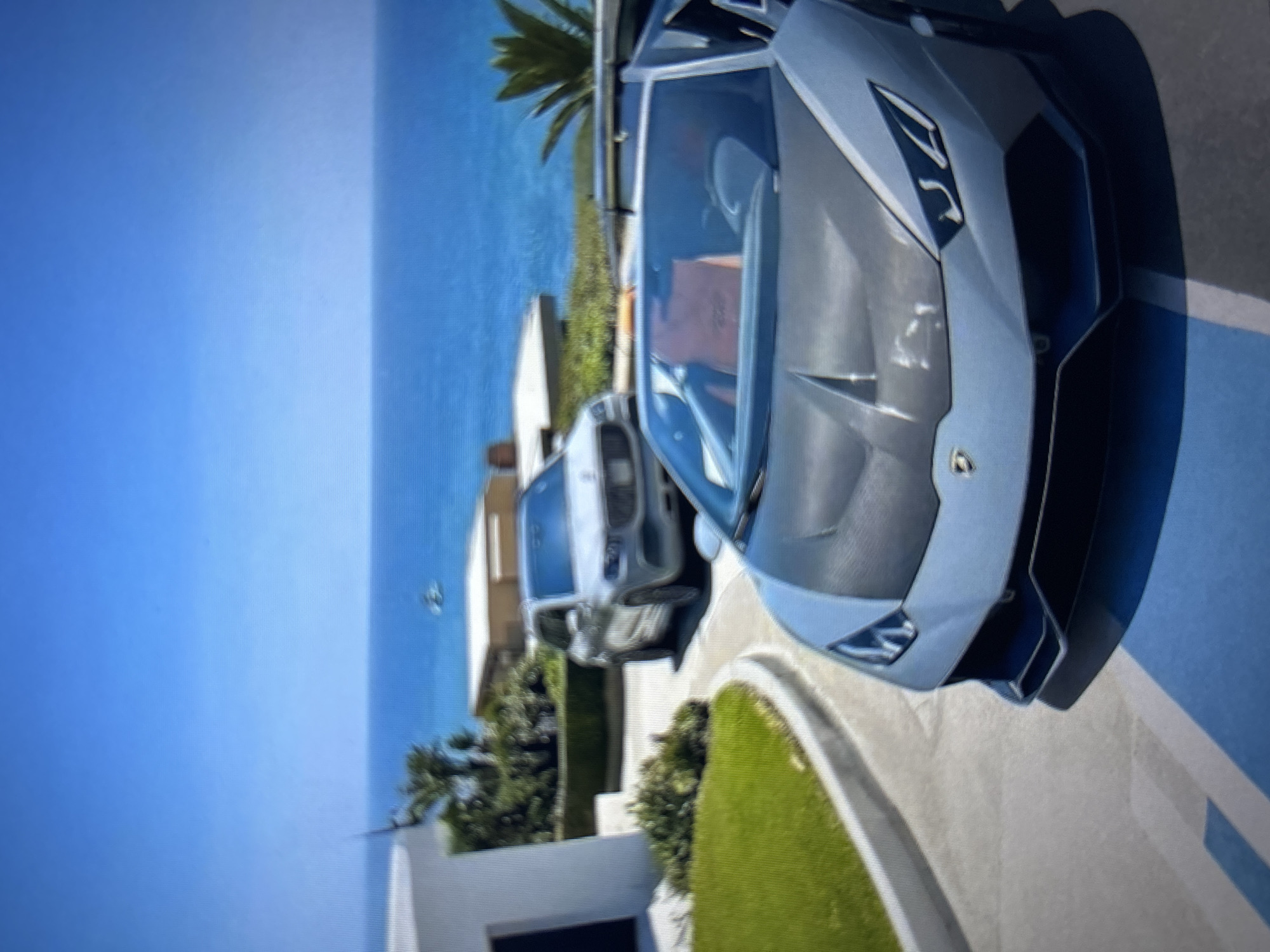 The image displays a sunlit coastal property in Burgas, Bulgaria. In the immediate foreground, a light grey Lamborghini Huracán Spyder, featuring a dark hood, is parked on a paved surface marked with blue and white lines, indicating a parking space. The vehicle's orange interior is visible through the windshield. Further back on a curving paved driveway, a light-colored Mercedes-Benz GLS-Class SUV is present, appearing to be in motion or parked. The driveway is bordered by a well-maintained green lawn and various plants, including palm trees and manicured hedges. A segment of a white, modern building is visible on the left. The background features a clear blue sky transitioning to a deep blue ocean, where a single white boat or yacht is discernible in the distance. The scene is illuminated by bright daylight, and no people are visible.