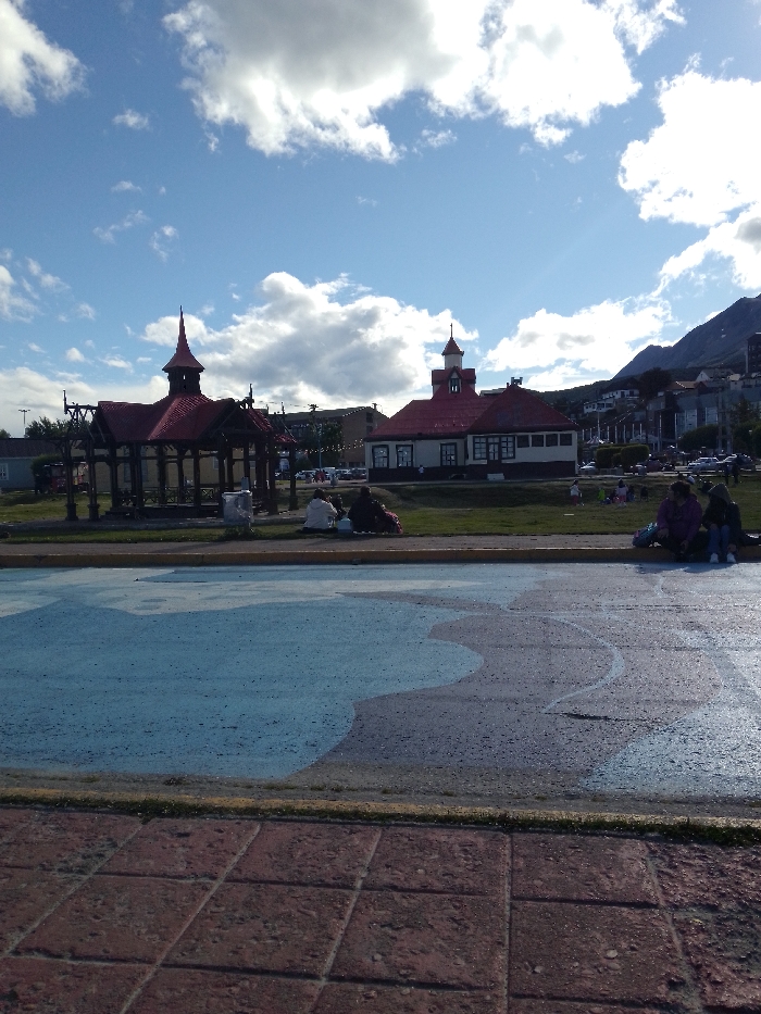 A view of Ushuaia, Argentina under a partly cloudy sky. In the foreground is a public square with a partially painted map on the ground. Behind it, a green strip of grass is lined with buildings, including a gazebo and a historic red-roofed building. Further back are trees, residential structures, and the foothills of the Martial Mountains. Several people sit or stand on the grass, enjoying the scenery.