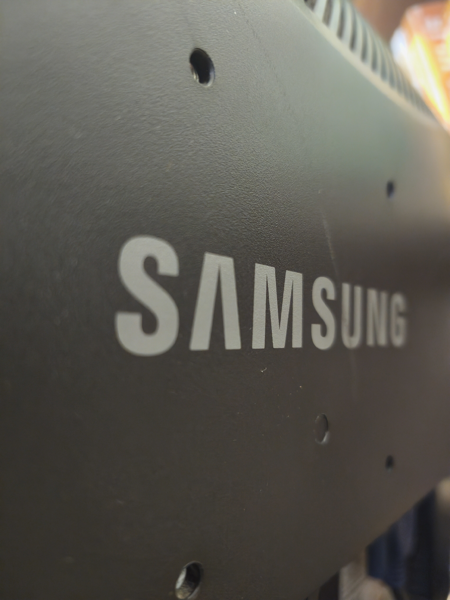 This media file is a close-up, angled shot of the back of a Samsung electronic device, likely a monitor or TV, in Kyiv, Ukraine. The surface is a dark, textured matte black. The prominent feature is the silver Samsung logo, clearly visible in the center of the frame, with the letters "SAM" on the left and "SUNG" on the right.
There are several small, circular indentations or holes scattered across the surface, suggestive of mounting points or ventilation. One such hole is particularly noticeable to the left of the logo. Towards the top right of the image, a series of ventilation slits are visible, indicating the rear of the device. The lighting suggests an indoor setting, with some warm, out-of-focus light sources visible in the background, possibly indicating other electronic devices or ambient lighting. No people are present, and the focus is solely on the product itself. The overall impression is one of a functional, perhaps slightly aged, piece of consumer electronics.
