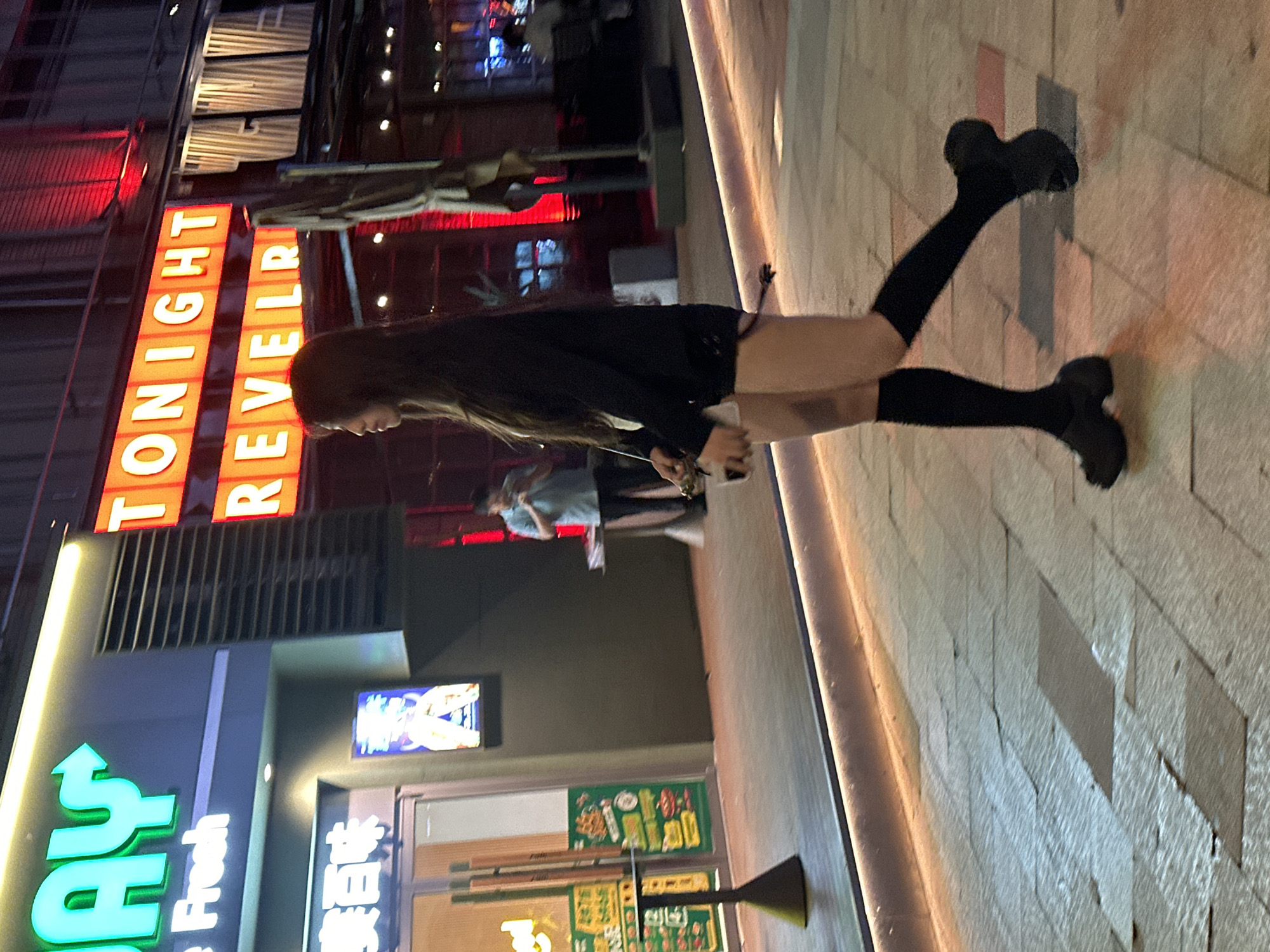 The image captures a street scene at night, likely in the Chongqing High-tech Zone, China, illuminated by neon and ambient lighting. In the foreground, a young woman with long dark hair is captured in profile, walking from right to left. She is wearing a black long-sleeved top, black shorts, black knee-high socks, and chunky black platform sandals. She is carrying a small handbag. Behind her, a man in a light blue short-sleeved shirt and dark pants stands near a table, holding a phone to his ear. Further in the background, neon signs display text, including "AY" in bright green and "TONIGHT REVELRY" in orange. Storefronts with glass doors are visible, one with a menu board displaying food items. The ground is paved with large square tiles.