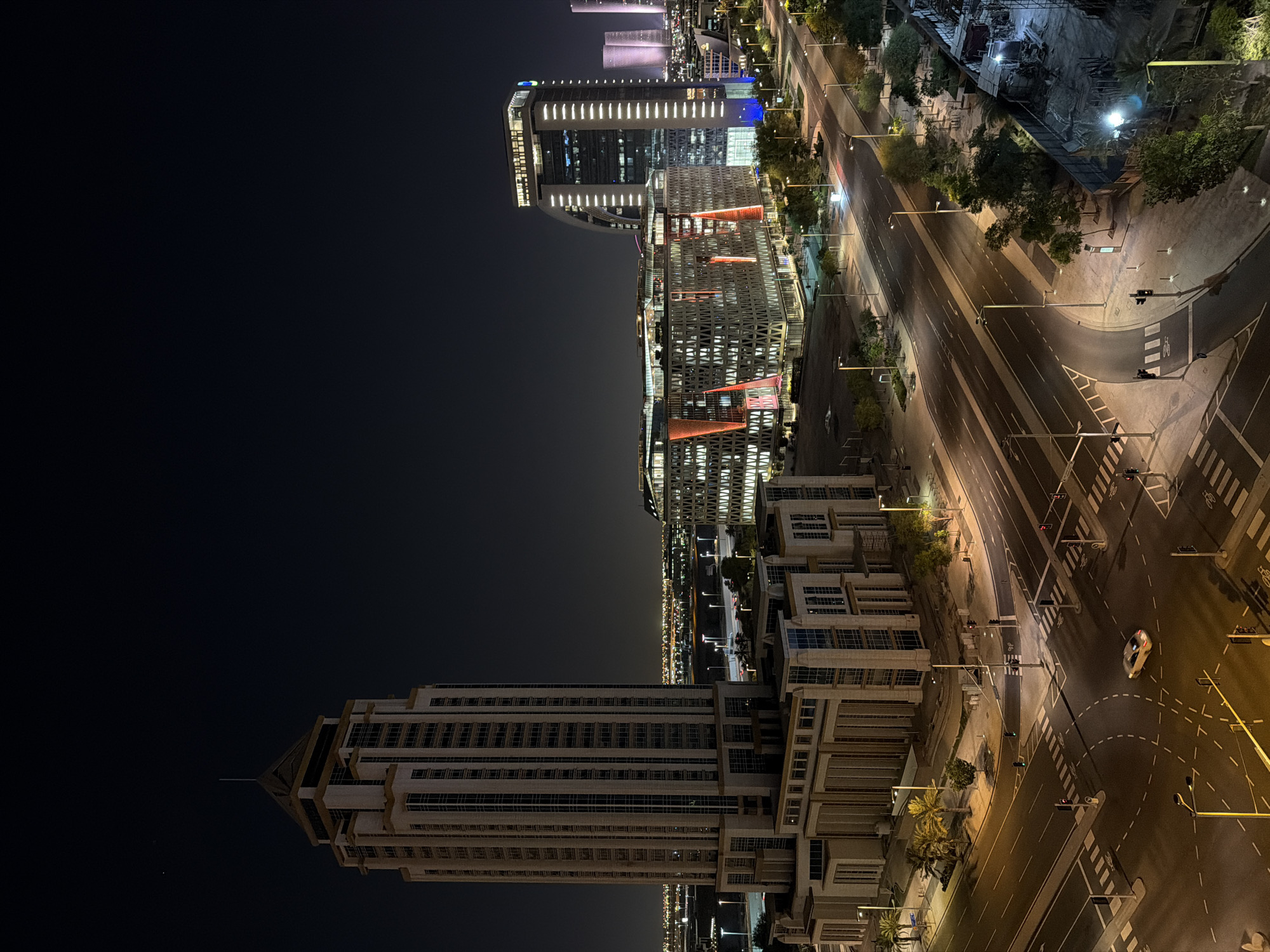 The image presents a high-angle, panoramic view of a modern cityscape at night, set against a dark, clear sky, consistent with an urban environment like Lusail, Qatar.
In the foreground, a large, multi-story building with a distinctive, tapering design and numerous windows rises prominently on the left. Its light-colored facade is illuminated by ambient city lights. To its right, a cluster of sleek, modern skyscrapers dominates the middle ground. One notable building features prominent vertical strip lighting, while others exhibit intricate, geometric patterns of illumination and splashes of red and blue light. A small, multi-colored corporate logo is visible on one of these buildings.
Below the buildings, a wide, multi-lane road system weaves through the urban landscape. The roads are well-lit by numerous streetlights emitting a warm glow and appear largely empty, suggesting a late hour. Clear road markings, including lane dividers and pedestrian crossings, are visible, along with traffic lights at various intersections. A single light-colored car is captured in motion on one of these intersections.
Patches of green trees and landscaping are interspersed along the roads and between structures. In the upper right background, a construction site with temporary lighting indicates ongoing development. The overall impression is one of a clean, modern, and relatively quiet city at night. No people are discernible, and the weather appears calm and clear.