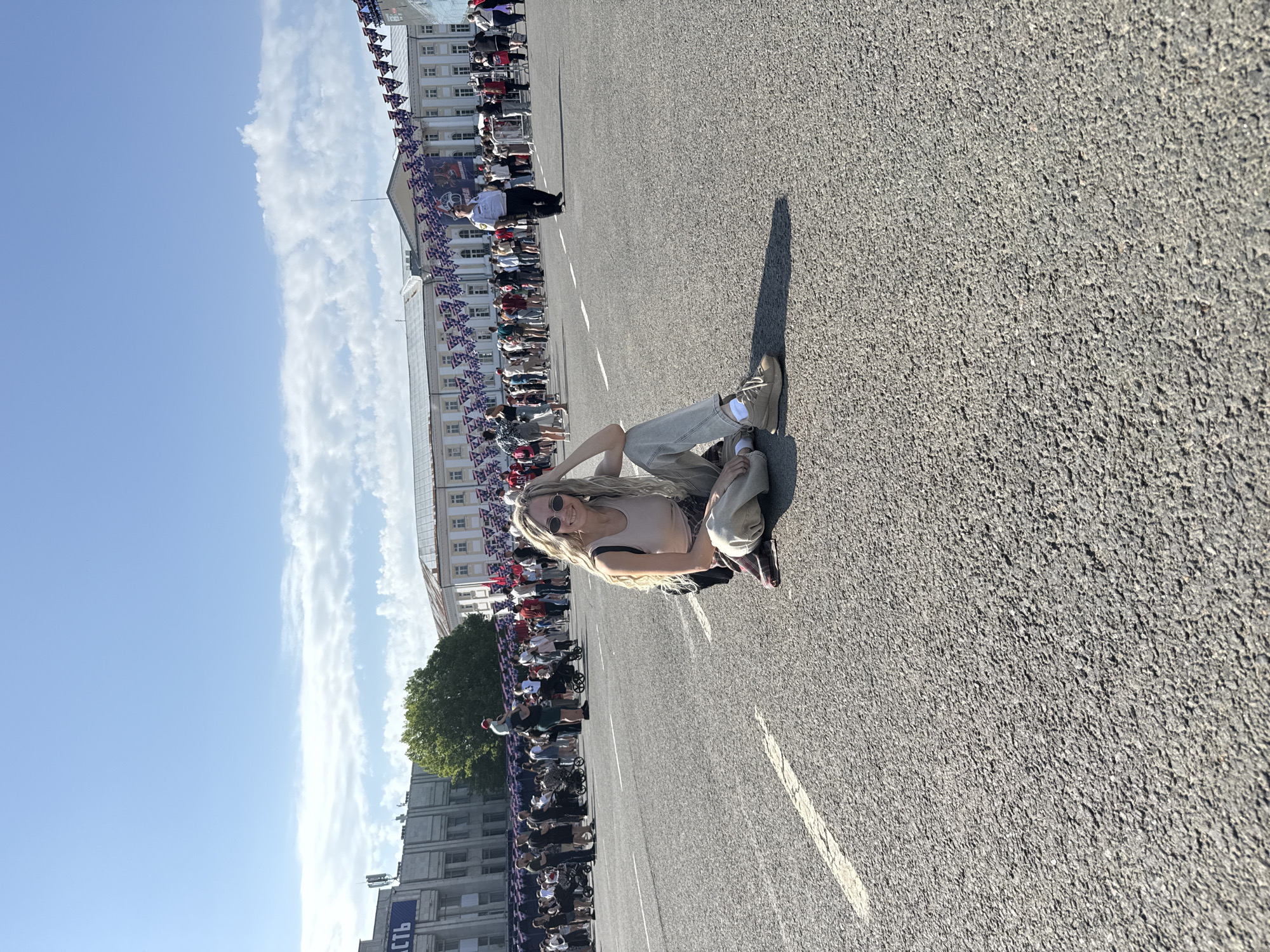 The image captures a person sitting on an asphalt surface, likely a street or plaza, in Заволжье, Russia. The person, with blonde hair and sunglasses, is wearing a light-colored top and pants. Behind the person, a large crowd is gathered along both sides of the street, potentially for a parade or public event. Banners or flags are lining the buildings, suggesting a festive atmosphere. In the background, there are buildings of varying heights. The sky is blue with some clouds.