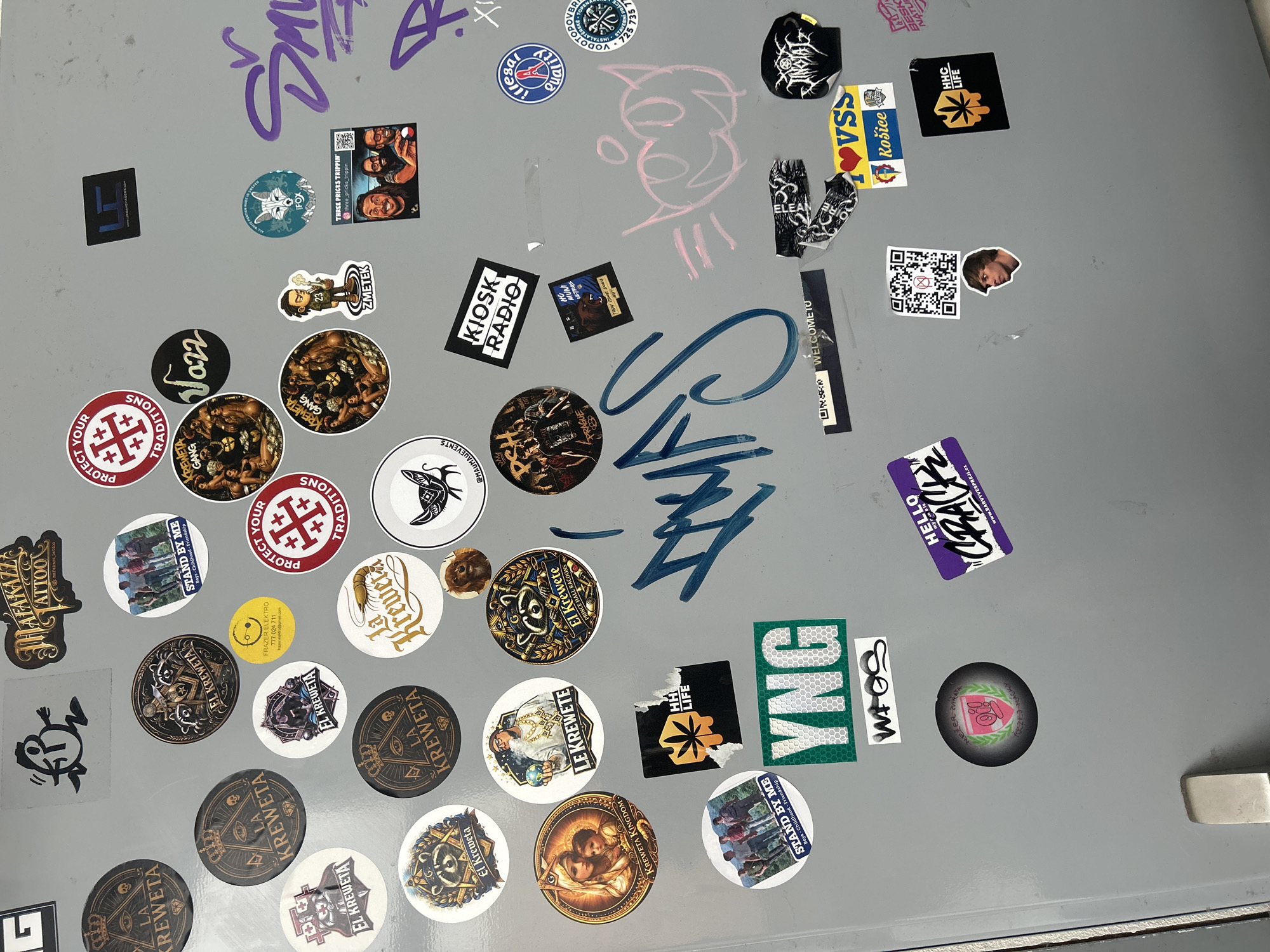 This is a medium-shot, eye-level, full-frame photograph of a collection of stickers on a gray surface, likely a refrigerator door, given the presence of a handle on the right side of the frame. The stickers are varied in shape, size, and design, featuring text, illustrations, and photographs.
Prominent subjects include stylized graphic designs, cartoon characters, and photographs of people. Some stickers have a tattoo or graffiti aesthetic, with bold lettering and intricate patterns. Several circular stickers feature the text "EL KREWETA" and "LA KREWETA" along with imagery of crowns and eyes, suggesting a group or brand affiliation. Another recurring motif is the word "KREWETA GANG".
There are also stickers with text like "KIOSK RADIO," "PROTECT YOUR TRADITIONS," "STAND BY ME," "HHC LIFE," and "YNG." One sticker displays a QR code. A few stickers have a vintage or retro feel, while others appear more modern. The overall impression is that of a personal collection of stickers, possibly accumulated from various sources or representing personal interests and affiliations. The lighting is even, indicating an indoor setting, and there are no obvious weather cues. The image appears to be taken in Brno, Czechia, based on the presence of the word "Brno" on one of the stickers.