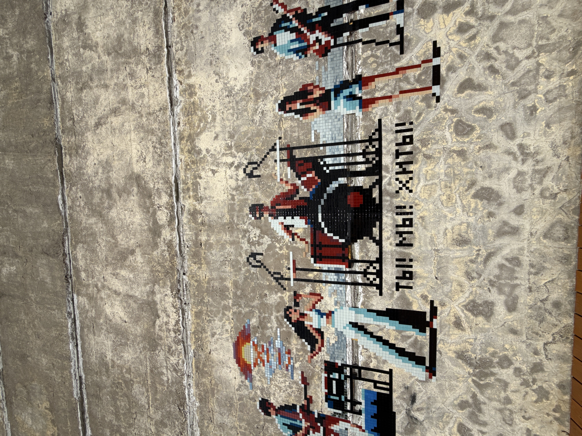 The image shows a mosaic on a concrete wall. The mosaic depicts a pixelated representation of people or figures standing in rows. The wall appears to be aged and weathered. A Russian inscription is visible, consisting of large pixelated letters. The location is Saint Petersburg, Russia.