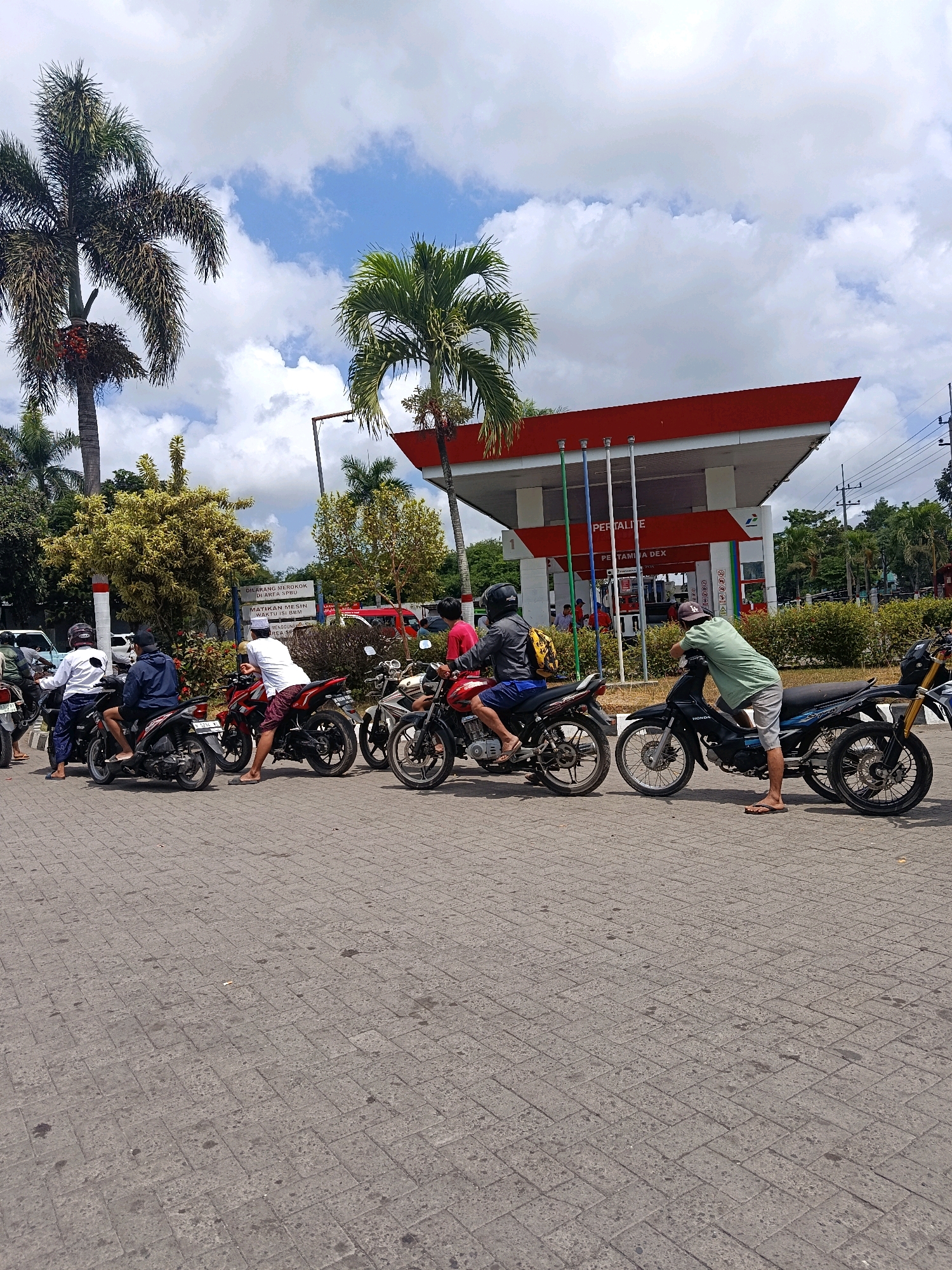 In Tempeh Lor, Indonesia, a queue of motorcycles with riders is forming at a Pertamina gas station. The station's canopy is painted red and white, with the Pertamina logo visible. Several palm trees are present, with lush green foliage and some bearing fruit. The sky is partly cloudy, with patches of blue visible. The ground is paved with interlocking bricks, some of which show signs of wear and staining. The riders appear to be waiting patiently, some are seated on their motorcycles, while others are standing or leaning. One person in a green shirt is leaning over a black motorcycle, while another person in a white shirt is standing next to a red motorcycle. A person wearing a white shirt and a dark blue jacket is seated on a motorcycle in the foreground. Another person in a white jacket is seated on a motorcycle further back. The scene is illuminated by daylight.