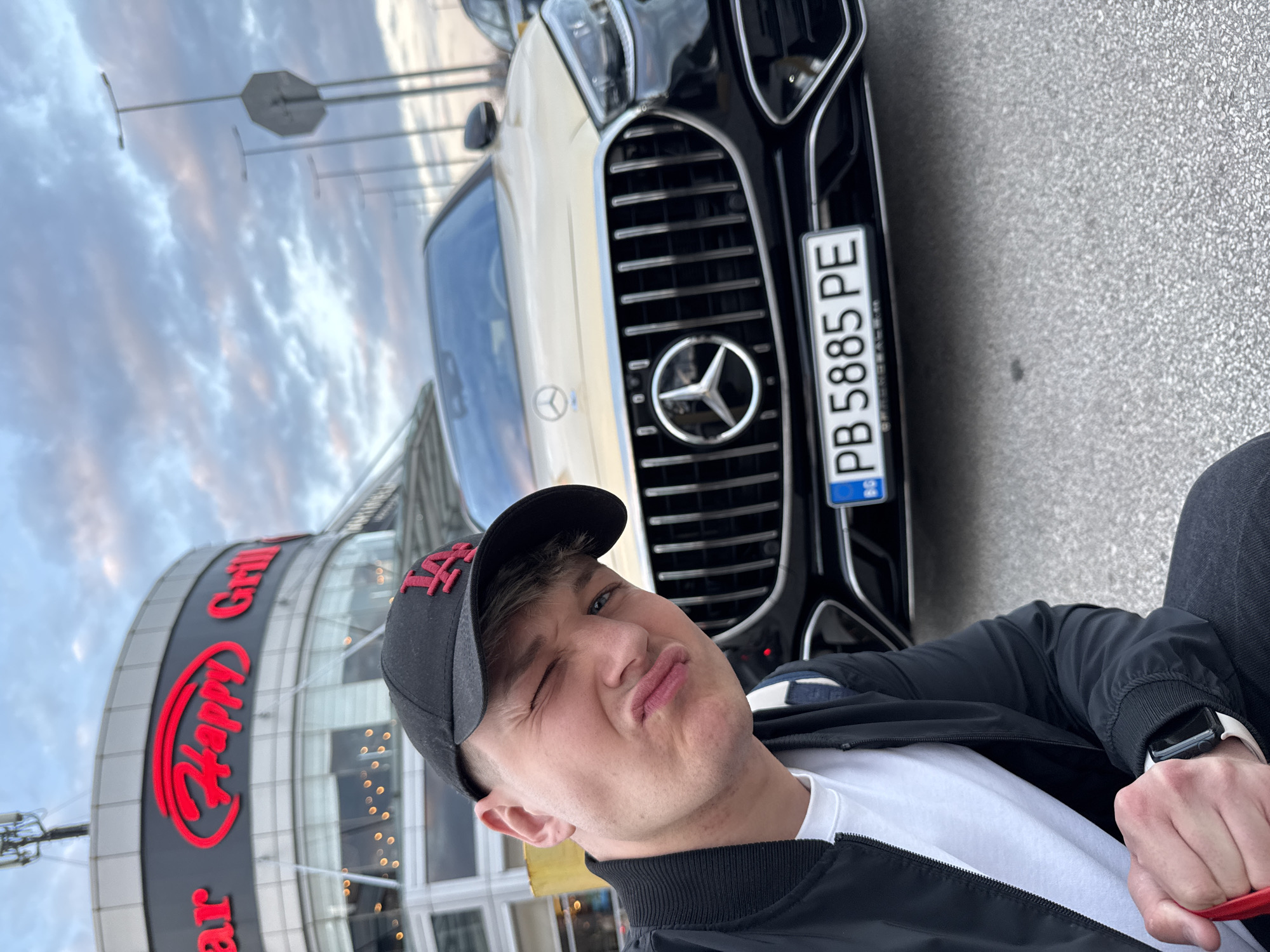 A young Caucasian male is depicted in a selfie, winking his left eye and pursing his lips, positioned in the lower-left foreground. He wears a black baseball cap with a red "LA" logo, a black bomber jacket, a white t-shirt, and dark pants. A white Apple Watch is visible on his left wrist. Behind him is a two-tone Mercedes-Benz sedan, featuring a gold/champagne upper body and a black lower section, with a prominent grille and the Mercedes-Benz emblem. The car displays a Bulgarian license plate, "PB 5885 PE", indicating registration in Plovdiv. In the background, a multi-story building with a large red neon sign reading "Happy Grill" is visible, identifying the location as a "Happy Grill" restaurant in the city of Plovdiv, Bulgaria. The scene is an outdoor parking area or street, under an overcast sky with scattered clouds, suggesting late afternoon or early evening lighting. Streetlights and a hexagonal street sign are present in the distant background. The ground is dark gray asphalt pavement.