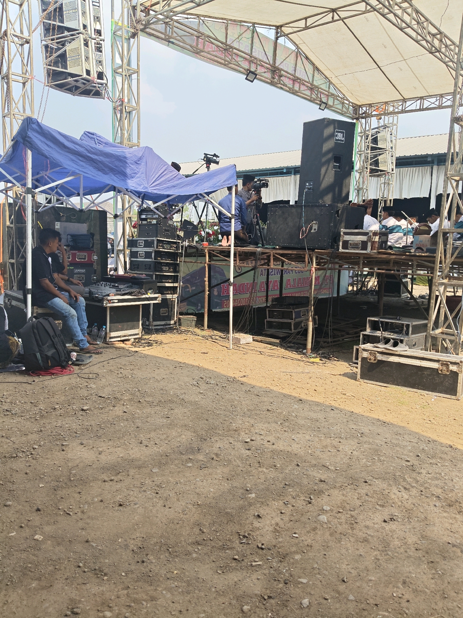 This is a medium shot, captured from a slightly elevated, forward-facing angle, of a stage setup in Bangsal, Indonesia. The stage is constructed from metal scaffolding, supporting a white tarp roof. The ground is dirt and gravel. A large black JBL speaker is prominently positioned on the right side of the stage. To the left, a man wearing a black t-shirt and blue jeans sits on a large black speaker case, operating a DJ mixing board. Two black backpacks and some red cabling are visible on the ground in front of him. A blue tarp is draped over a portion of the stage structure on the left. In the background, several people, some wearing white shirts, are present on the stage, and there are more speakers and audio equipment stacked on the scaffolding. A banner with text is visible behind the DJ setup. The sky above is clear and blue.