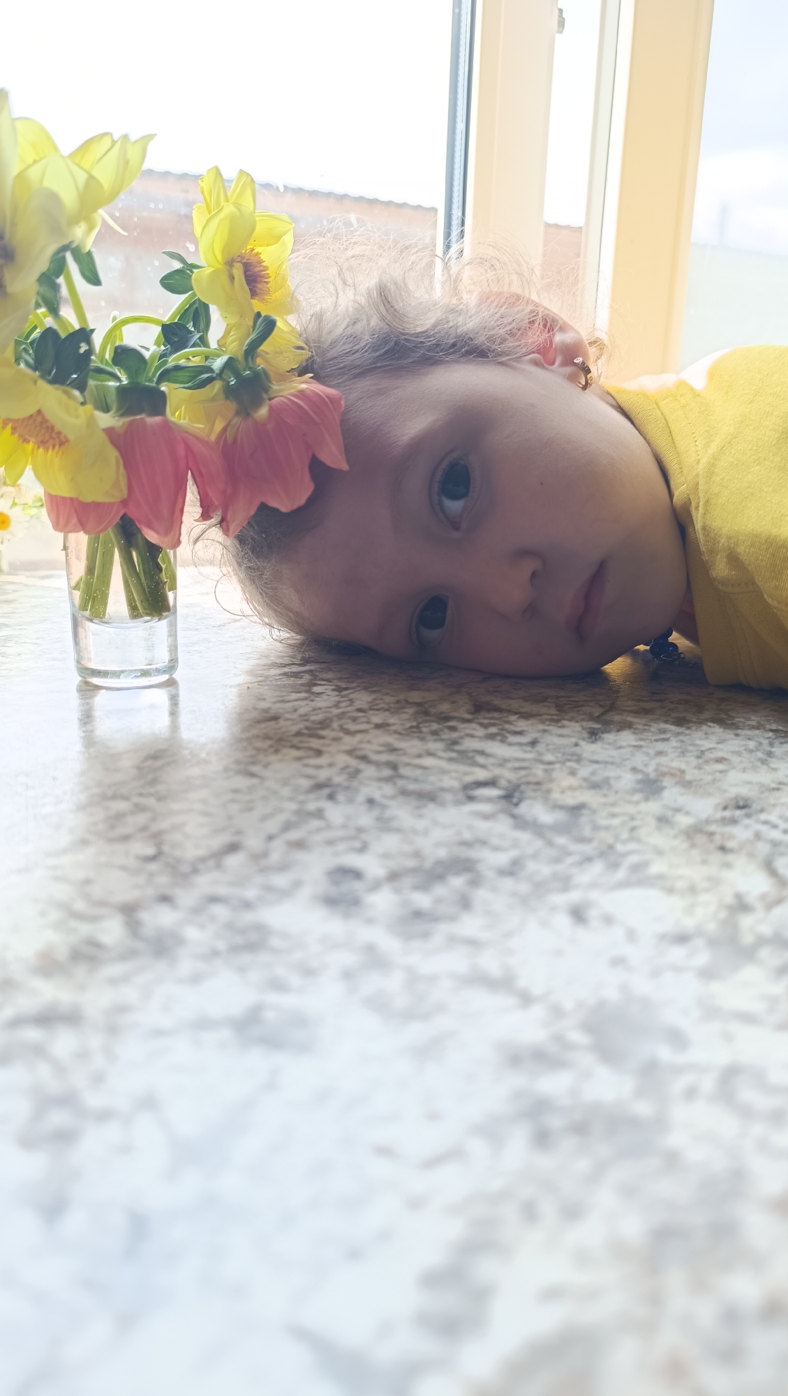 A child with blonde curly hair lies on a speckled countertop, their head resting on their left cheek. They are wearing a yellow shirt and have a slight smile. A small glass vase with yellow and pink flowers sits to their left. The background features a window with a light-colored frame and a hazy view of what appears to be buildings. The image is lit by natural light coming from the window.