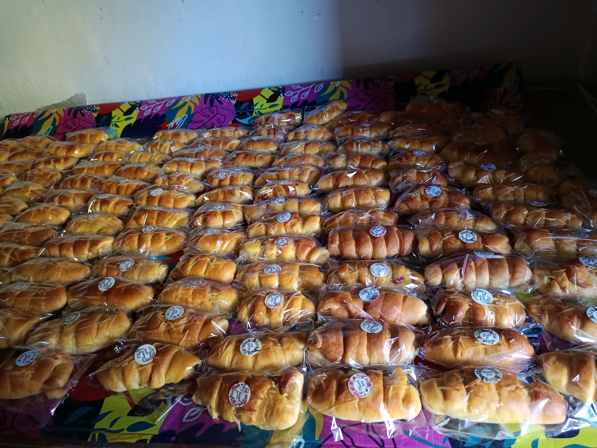 A table is covered with a brightly colored, patterned cloth. On top of the cloth, there are numerous individually wrapped loaves of bread. The loaves are golden brown and appear to be freshly baked. Many of them have a sprinkle of grated cheese on top. Each loaf is enclosed in clear plastic packaging, and many of these packages feature a circular sticker with floral designs and the words "Thank You" printed on them. The loaves are arranged in rows, filling the visible surface of the table. The background shows a white wall and a dark area, suggesting an indoor setting. The scene is likely a domestic or small commercial setting in Gunung Sekar, Indonesia, where these baked goods are prepared or displayed for sale or distribution.