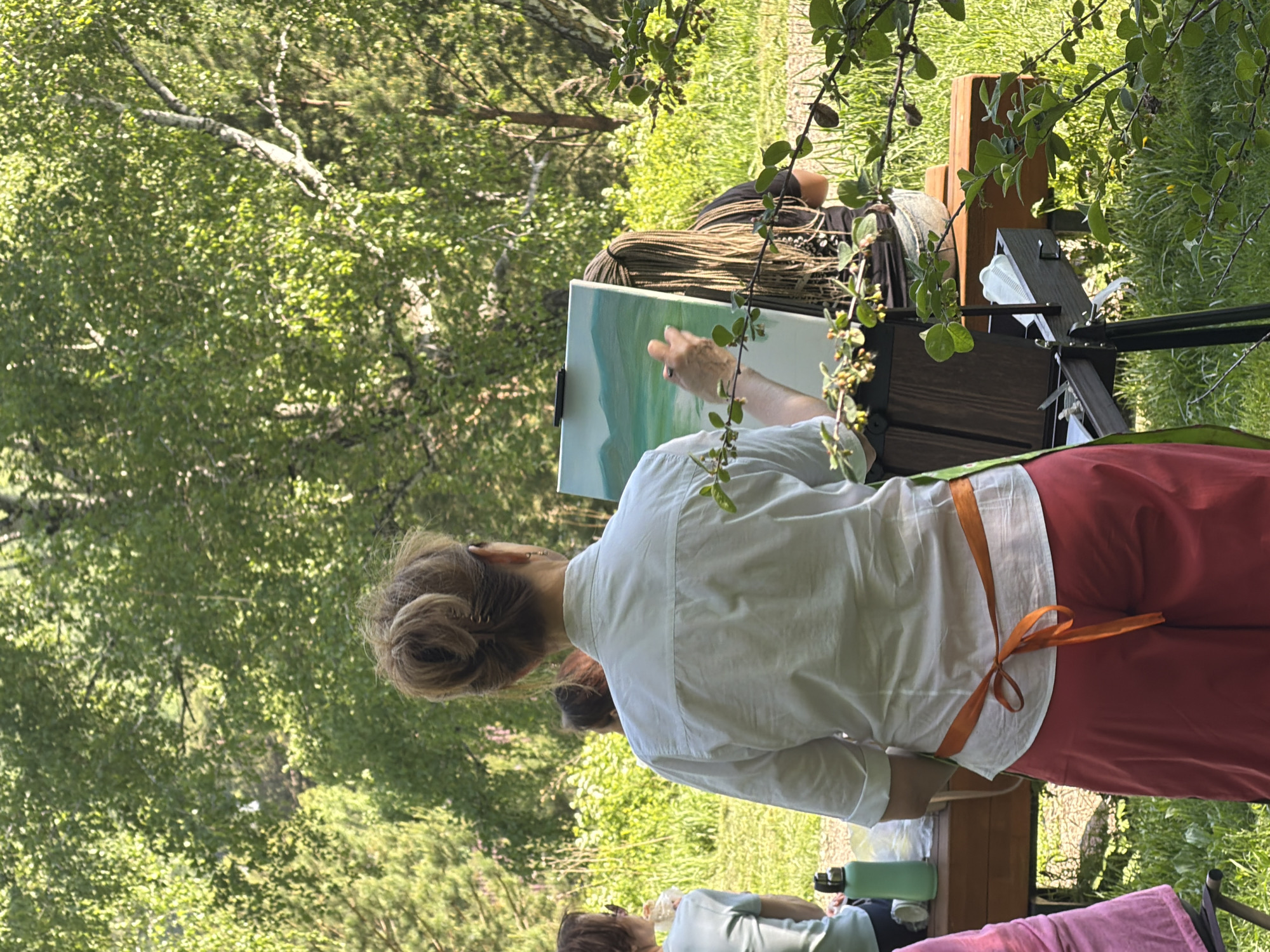 This image captures an outdoor plein air painting session in a lush, green environment, likely a park or natural area in Krasnoyarsk, Russia, under bright daylight. The main subject, viewed from behind, is a person with light brown hair styled in a bun, wearing a white long-sleeved shirt and red-orange pants with an orange apron or tie around their waist. They are actively painting a landscape scene on a canvas, which is mounted on an easel. The painting itself shows a calm, light blue sky over rolling green hills or mountains, reflecting the natural surroundings.
The background is rich with verdant foliage, including trees with pale bark (possibly birch) and various green bushes and grass, suggesting a vibrant natural setting. The bright illumination indicates a sunny day, likely during the morning or afternoon. Several other individuals are partially visible, suggesting a group activity. One person in the mid-ground has long, dark braided hair, and another's back and head are visible further away, along with a light green water bottle on a wooden surface. The scene conveys a peaceful and focused atmosphere, centered around artistic expression outdoors. There is no visible text in the image.